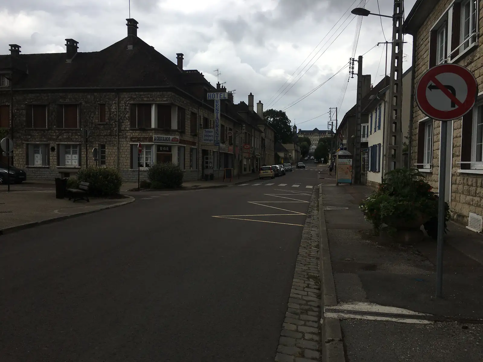 A small-town street on the Via Francigena lined with stone buildings, including a hotel-restaurant with an awning, under a darkening evening sky.