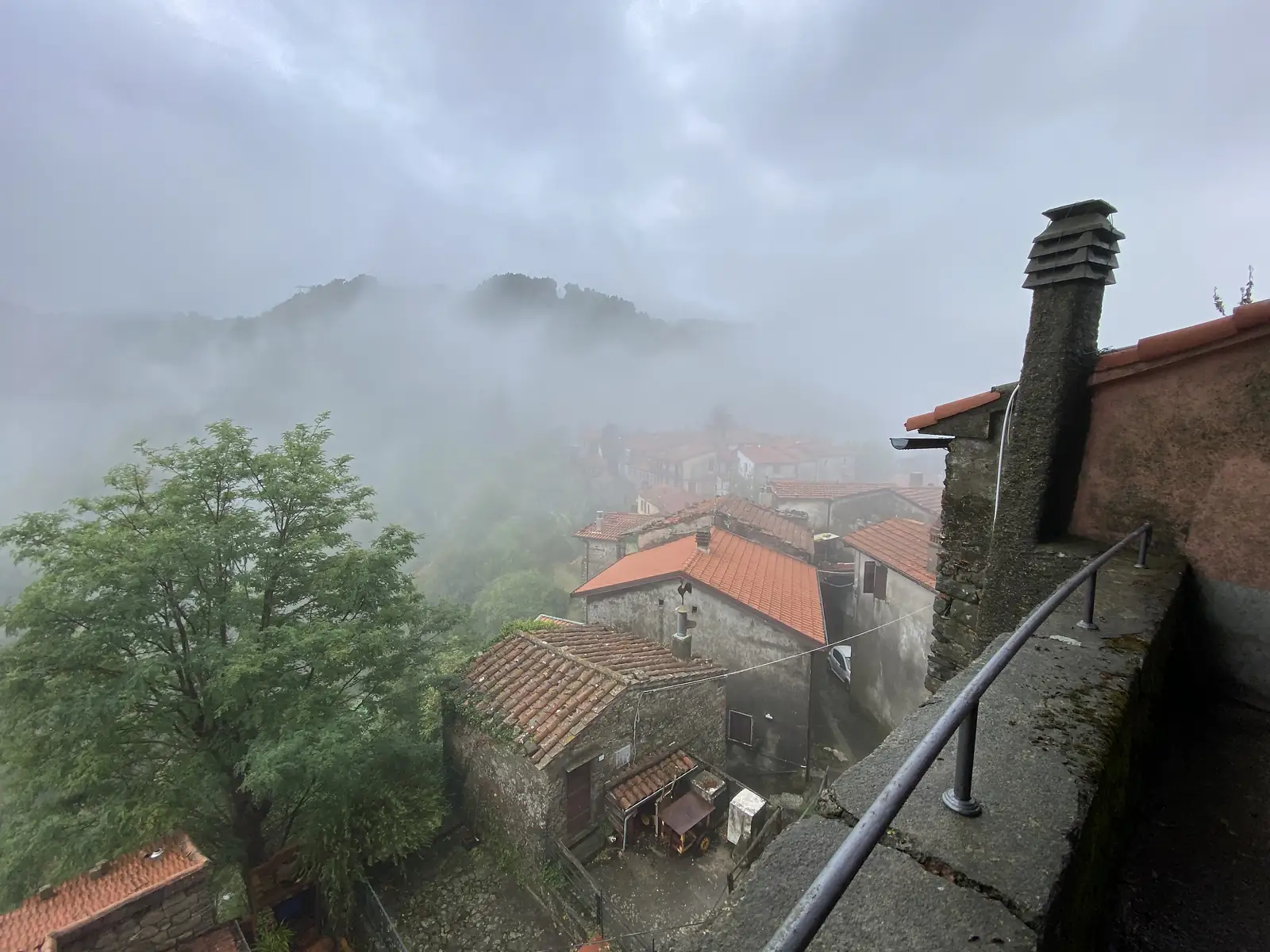 A misty hilltop village of terracotta-roofed stone houses, with a weathervane cockerel on one rooftop, seen past a chimney and a tree from a raised terrace.