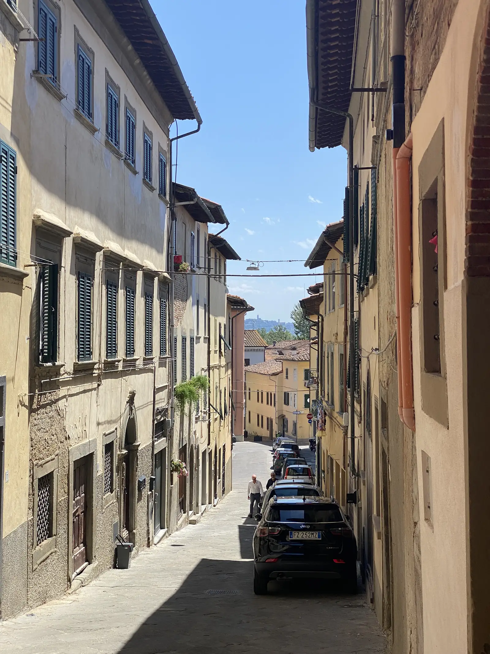 A narrow town street lined with tall pastel-coloured buildings and shuttered windows, cars parked along the cobbles and a pedestrian walking away from the camera.