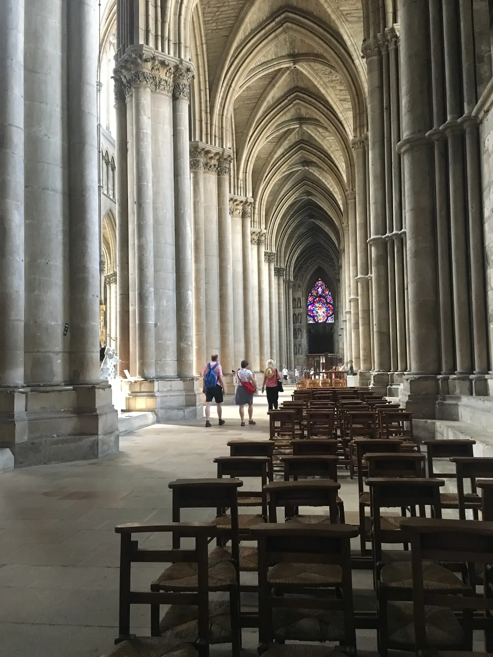 Rows of wooden chairs facing the transept of Reims Cathedral, with two rose windows of stained glass glowing above the crossing.
