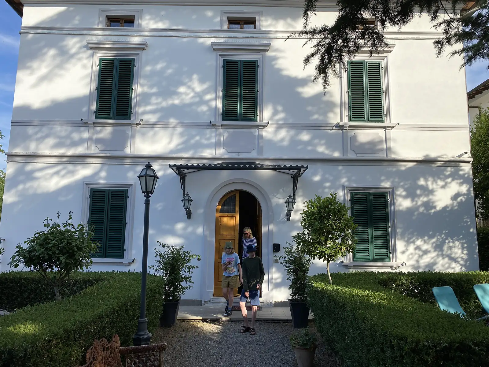 A family steps out through the arched wooden doorway of a white villa with green shuttered windows, past clipped hedges and potted trees in the garden.
