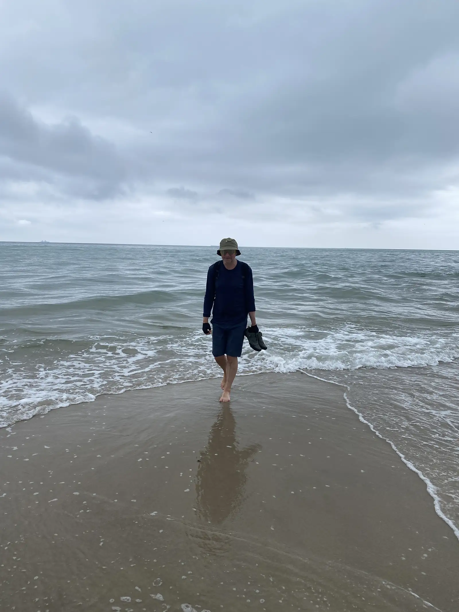 A person in a bucket hat wading barefoot out of the surf carrying their shoes.