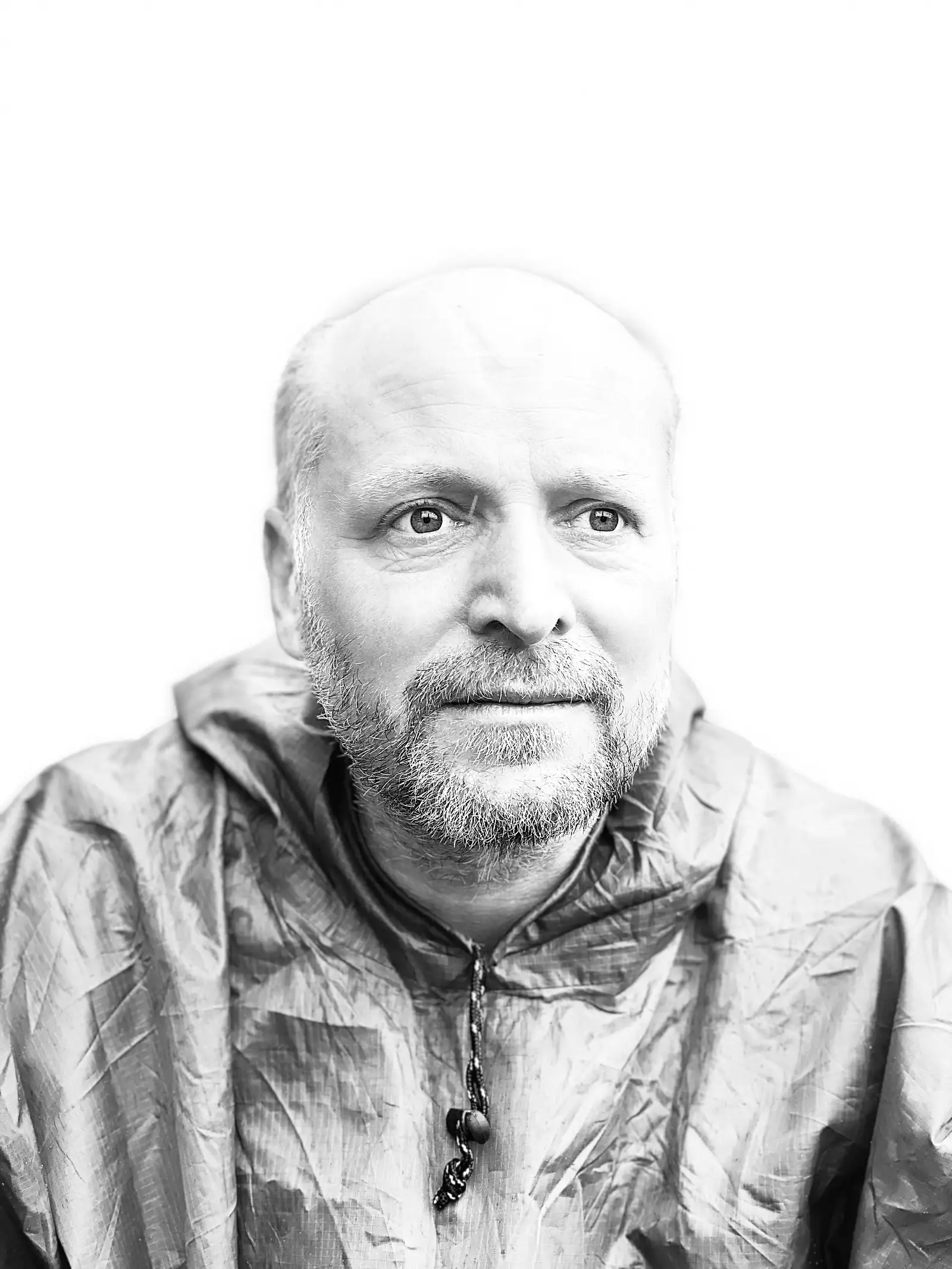 A bright high-key black-and-white portrait of a bearded person in a hooded waterproof, looking straight at the camera.