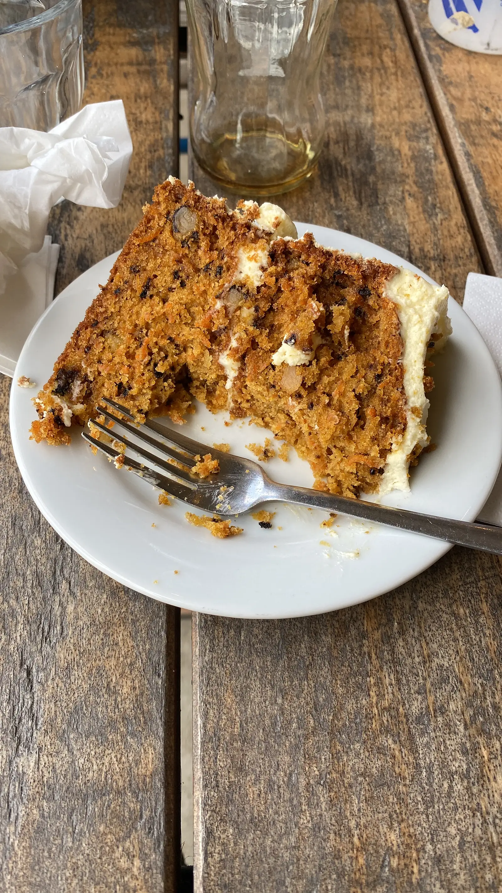 A thick slice of carrot cake with cream cheese icing on a white plate with a fork, on a weathered wooden table.
