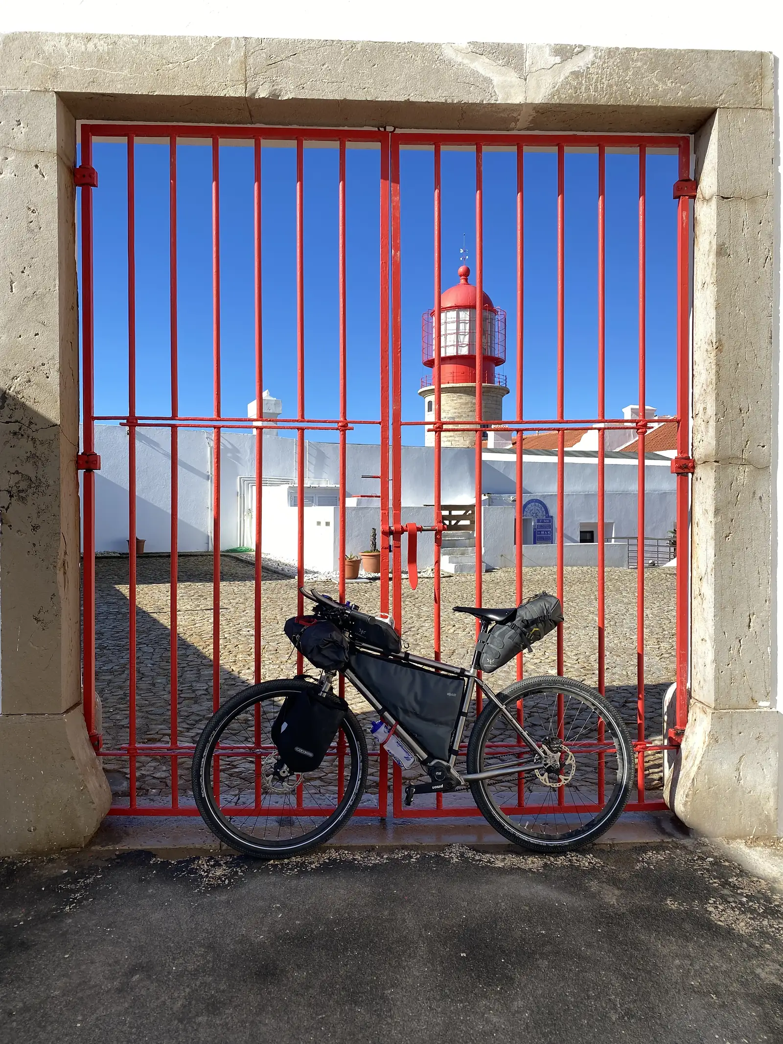 Loaded touring bike propped against tall red railings, framing a red-and-white striped lighthouse rising from a whitewashed courtyard under a cloudless blue sky.