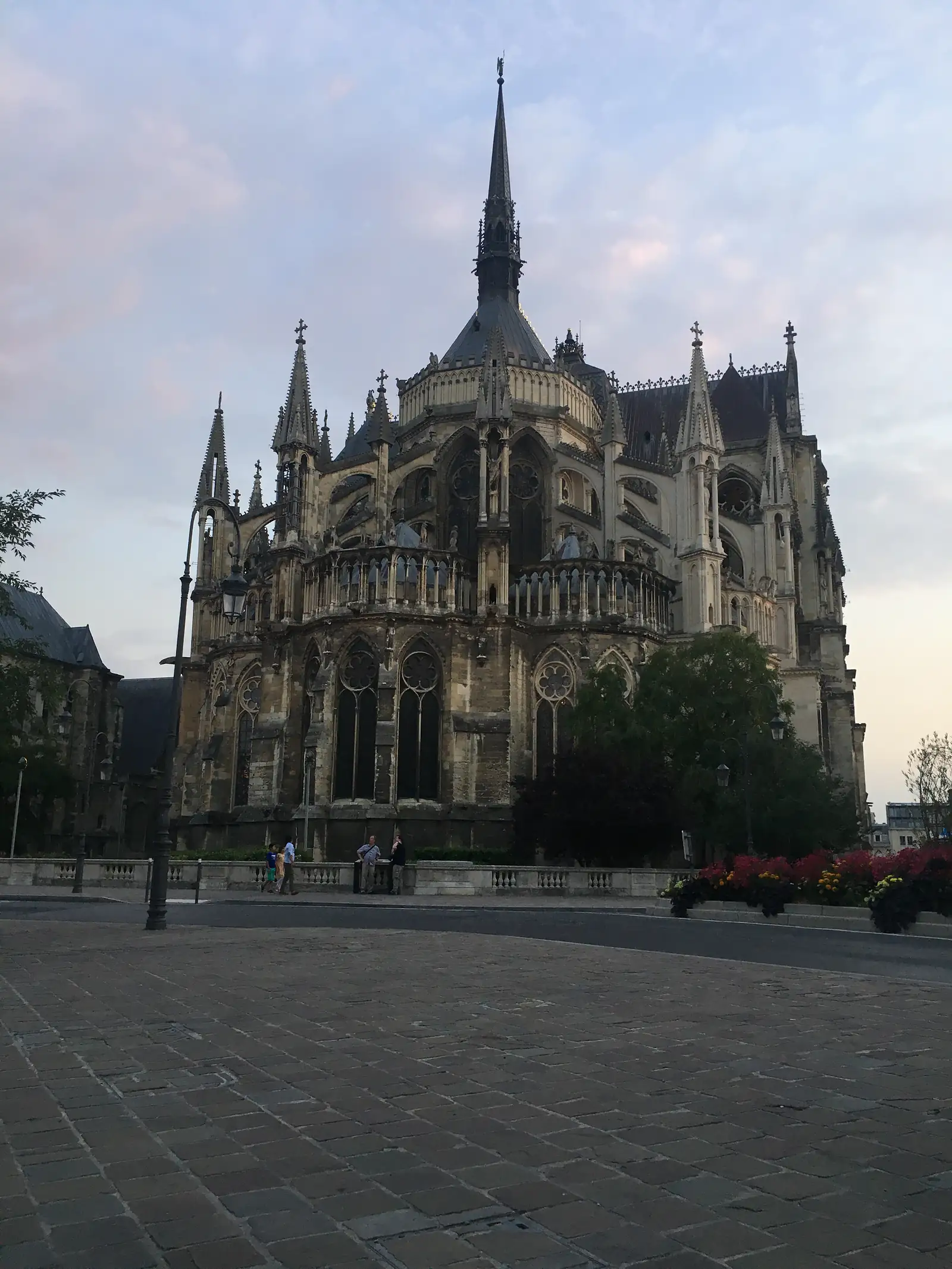 The soaring flèche and flying buttresses of Reims cathedral's east end rise above a hedge-lined pavement in fading evening light.