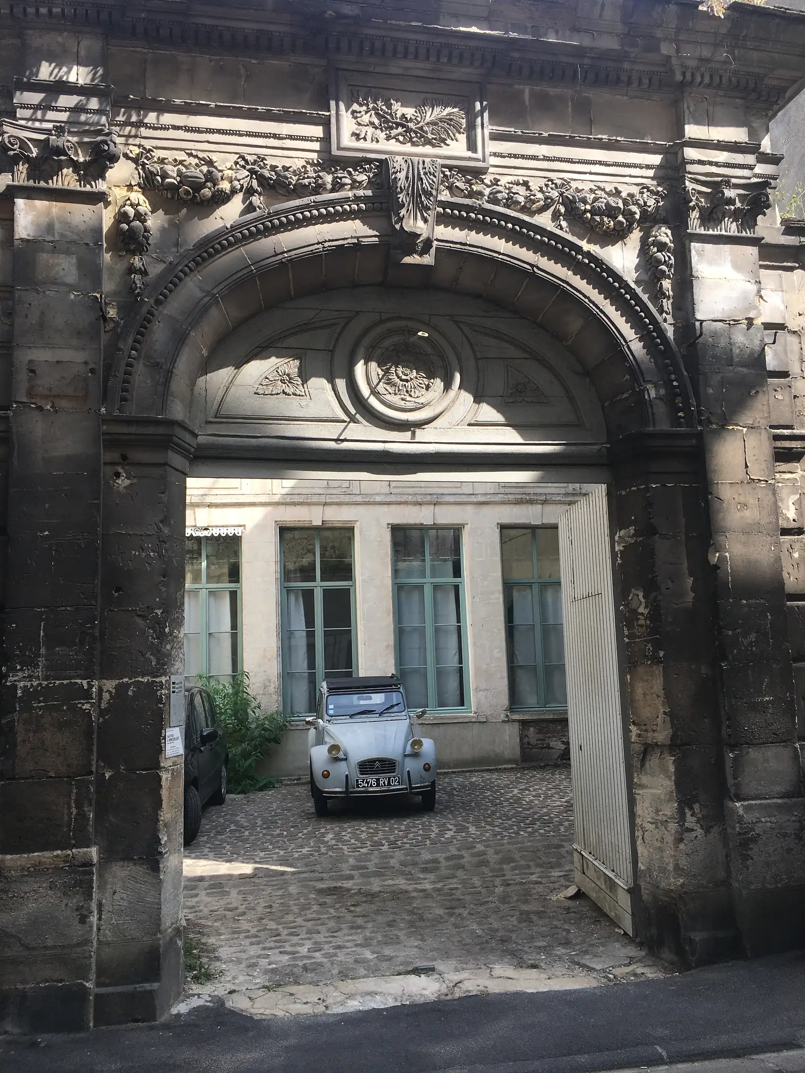 A weathered stone archway with carved floral decoration frames a courtyard where a vintage Citroën 2CV is parked.