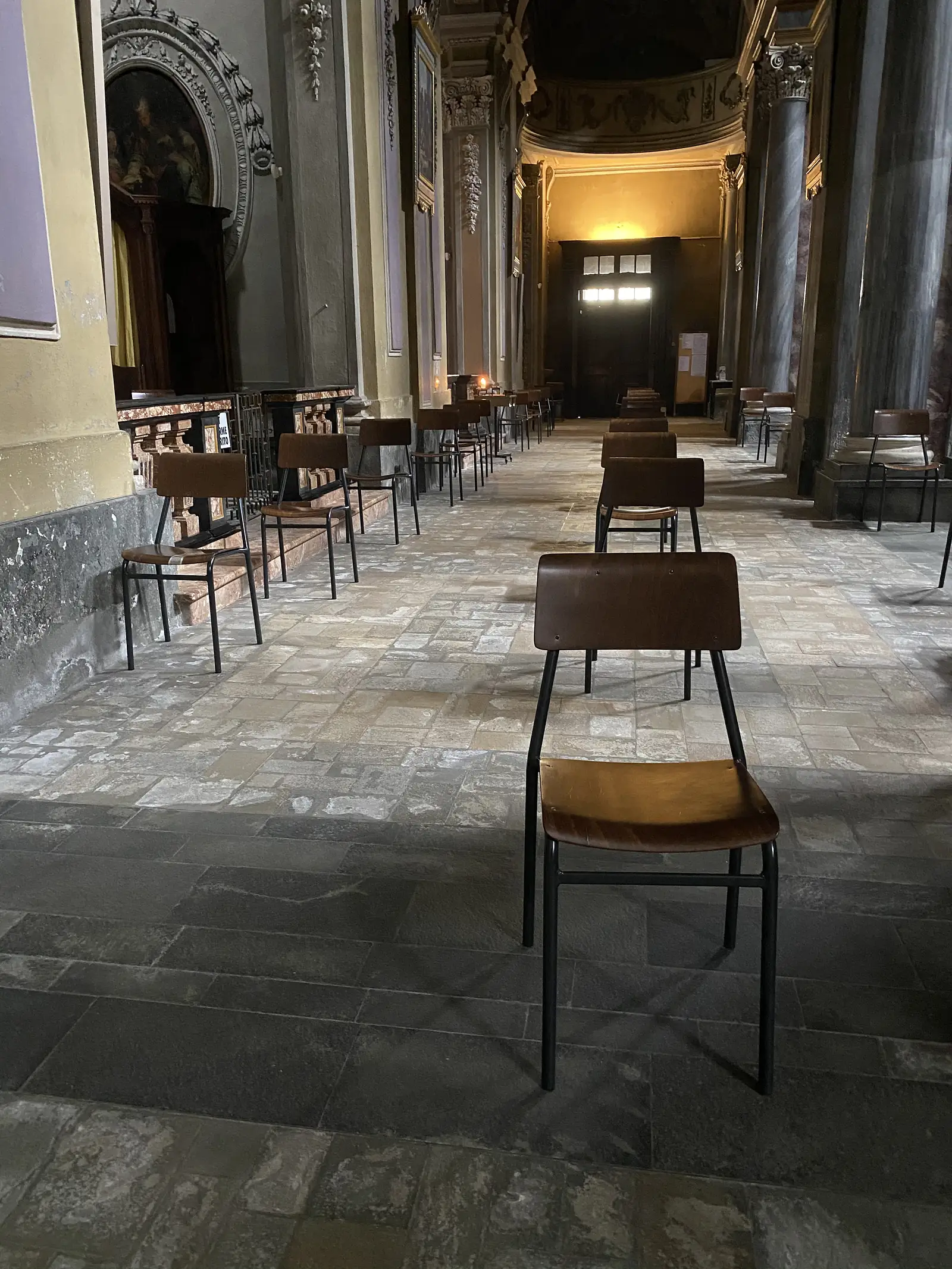 Rows of plain wooden chairs with black metal legs spaced along a stone-paved church aisle, leading towards a lit devotional shrine in the distance.
