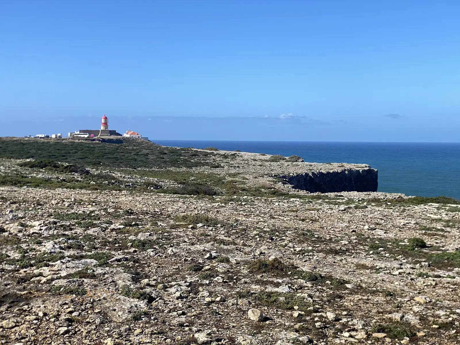 Flat, stony clifftop plateau of pale limestone and low green scrub running out to a sheer drop, with a small red-topped lighthouse and its white buildings on the point in the distance and deep blue sea beyond.