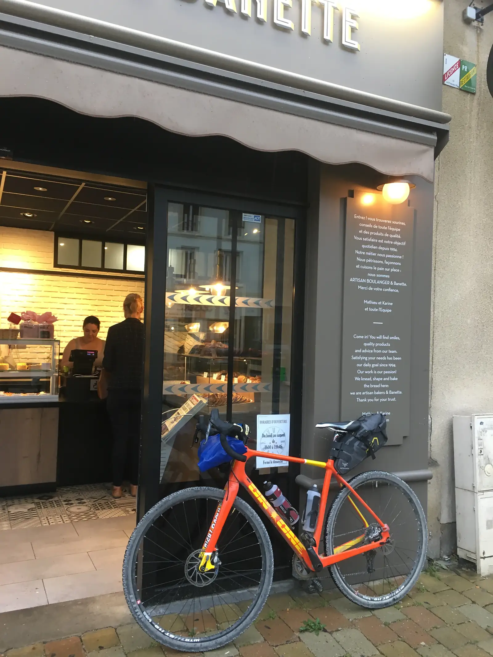 A mud-spattered orange gravel bike laden with bikepacking bags stands outside a bakery window, beneath a sign reading 'Come in! You will find smiles, quality products and advice from our team'.