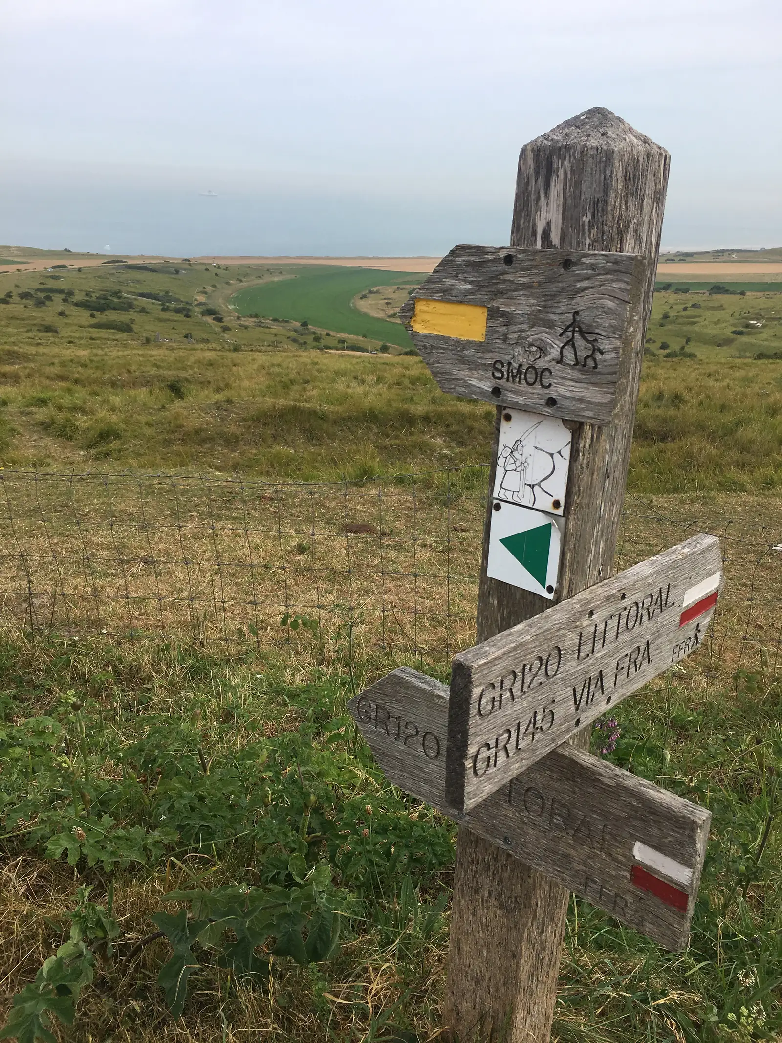 A wooden waymarker post carved with route names including GR120, GR145 Littoral, Via Francigena and Topal, painted with red-and-white trail blazes, overlooking the coast below.