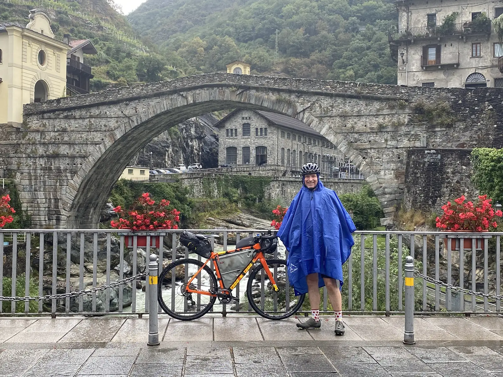A cyclist in a blue rain poncho and helmet smiles beside his loaded orange bike on a riverside promenade, with a high stone arch bridge and old buildings behind him.