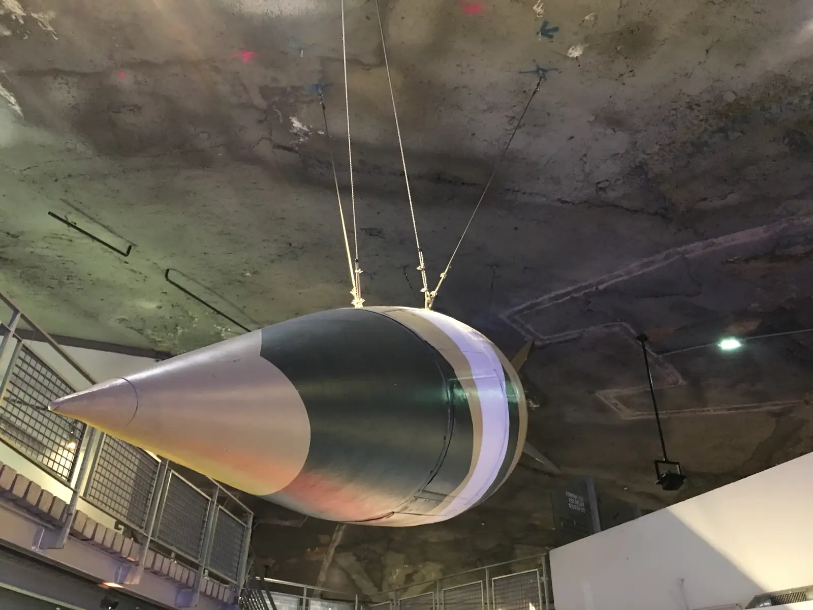 A grey-green missile replica with a sharply pointed nose hangs from wires beneath the tunnel's rough concrete ceiling.