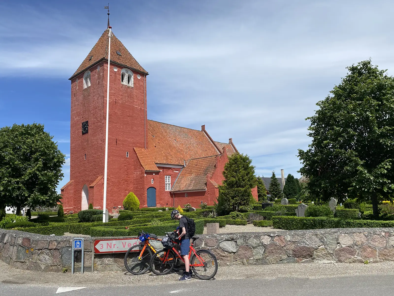 A child in a helmet standing with two loaded bikes on a road below a red-brick church with a tiled spire, beside a red cycle-route signpost.