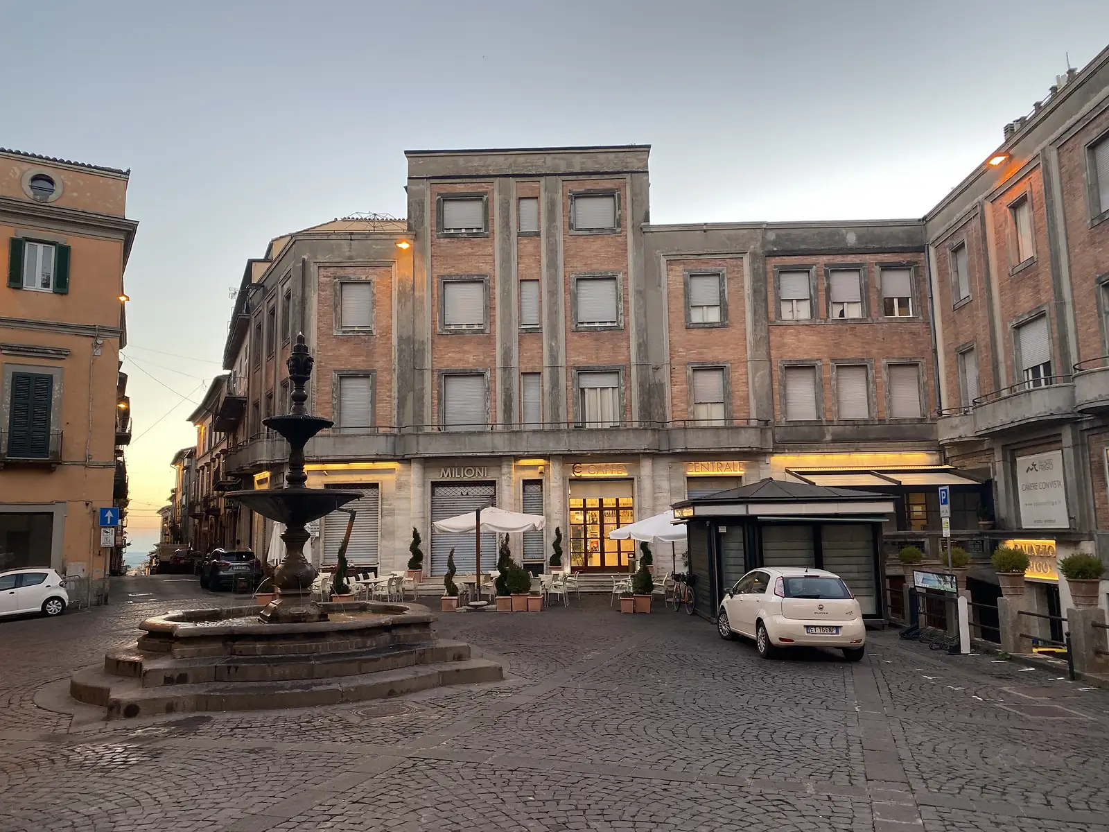 A stone fountain in a cobbled town square at dawn, with shuttered shopfronts and the "Caffè Centrale" sign lit beyond, cafe umbrellas and a parked car nearby.