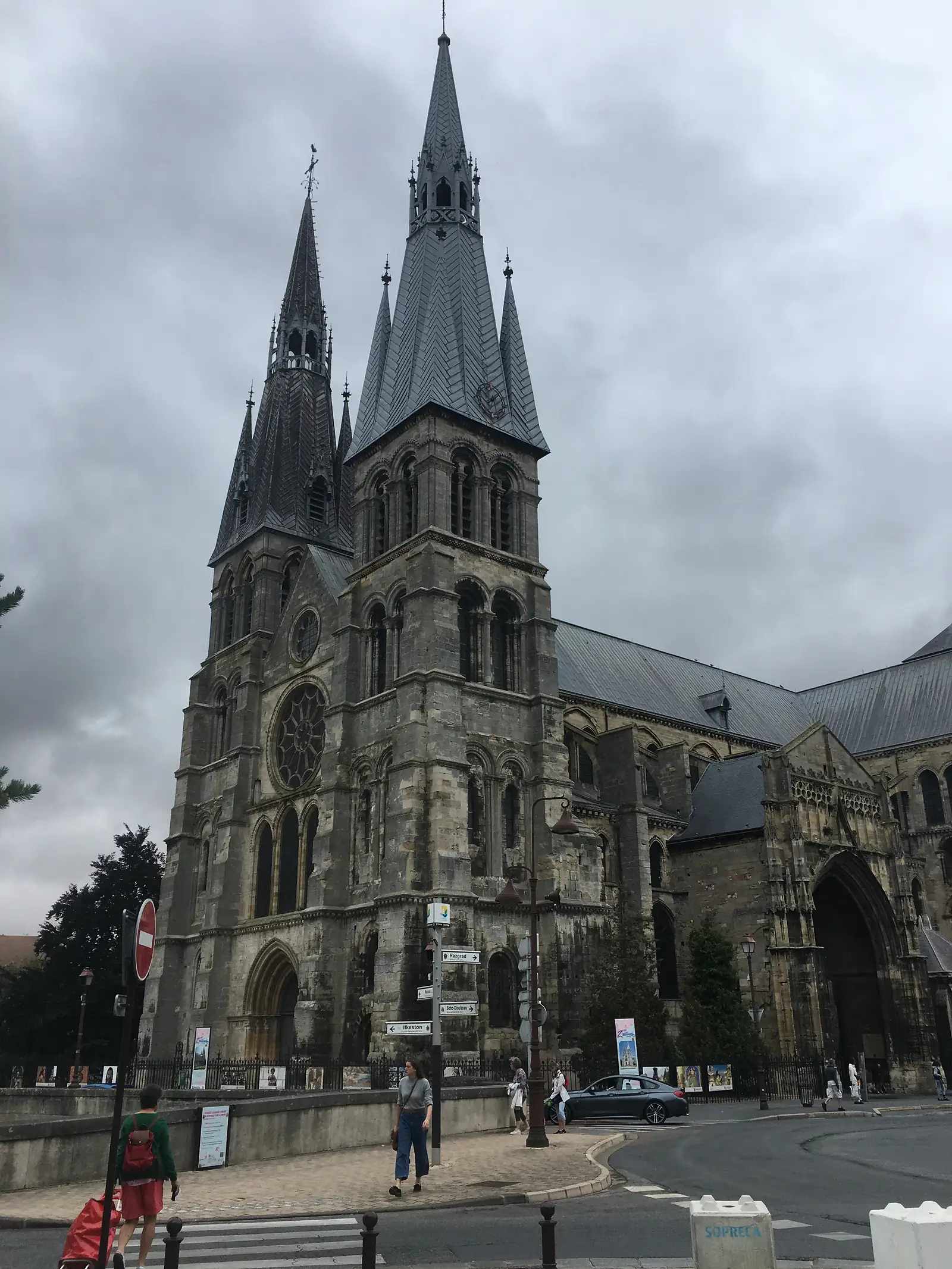 The twin spires and carved west front of a Gothic cathedral rise above a town square, with pedestrians and a directional signpost on the pavement below.