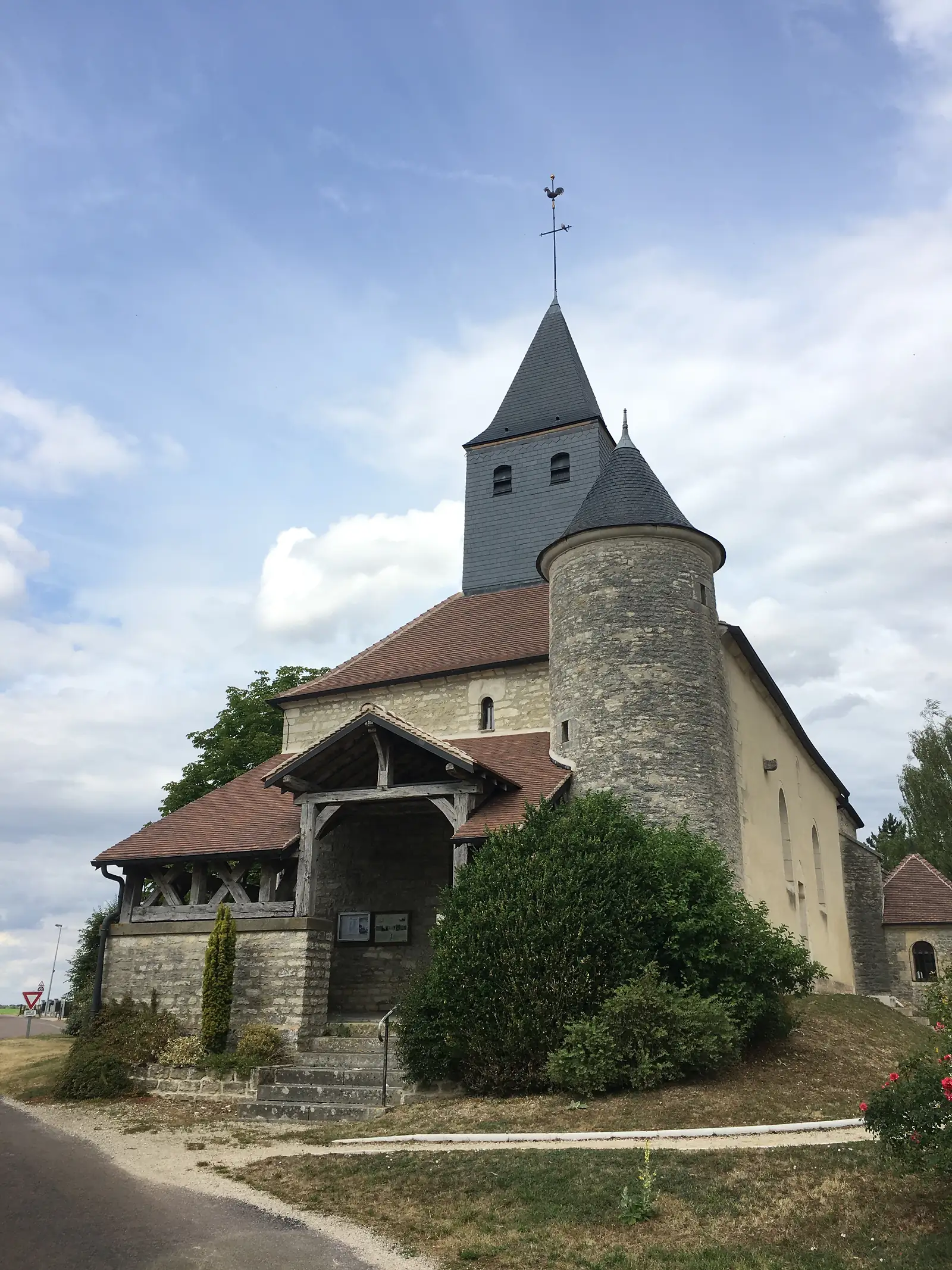 A church's round stone bell tower rises to a slate pyramidal spire topped with a weathervane, above a red-tiled nave roof.