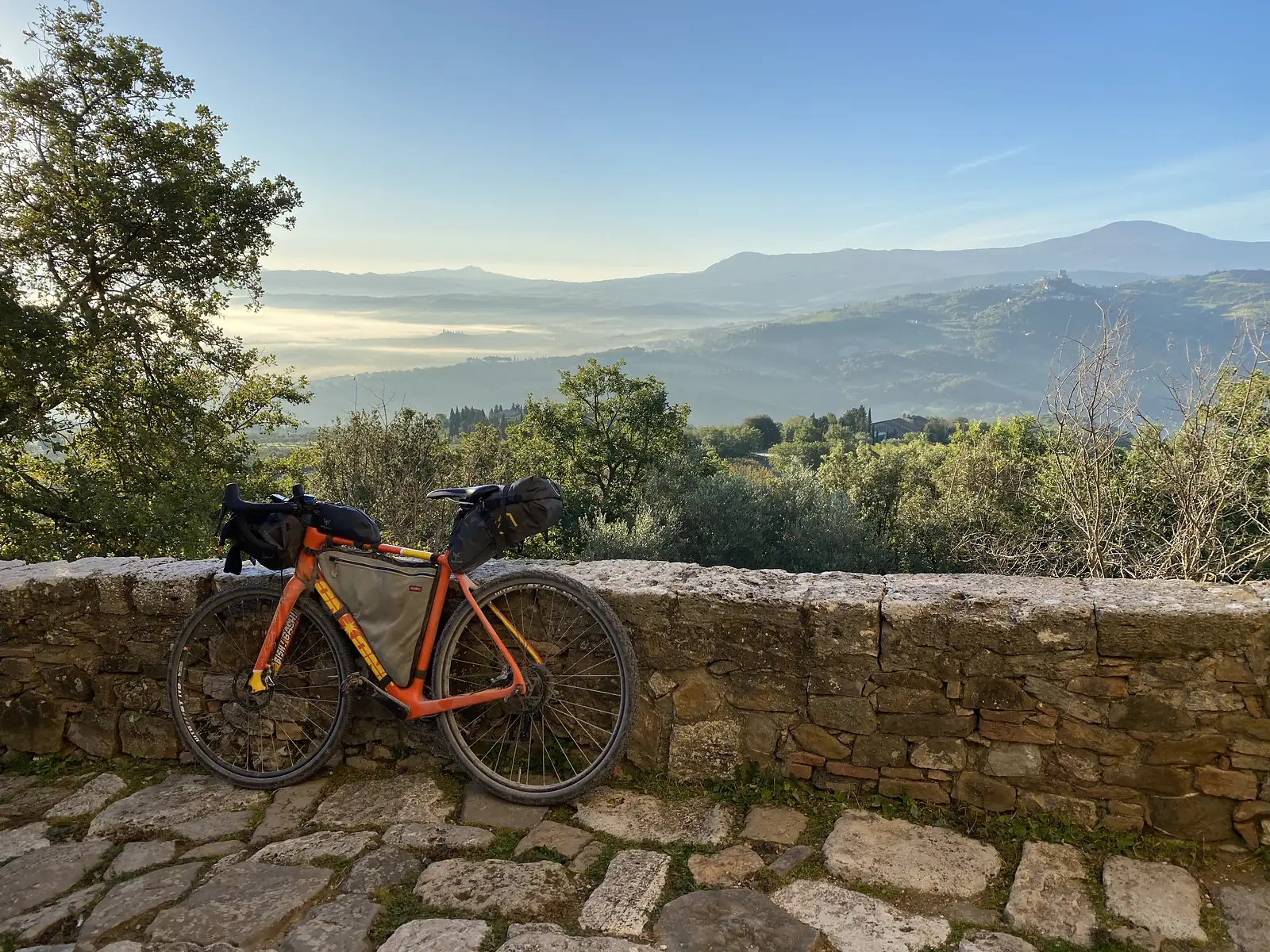 A loaded orange bikepacking bike leaning against a stone wall, overlooking a mist-filled valley with a hilltop town in the distance.
