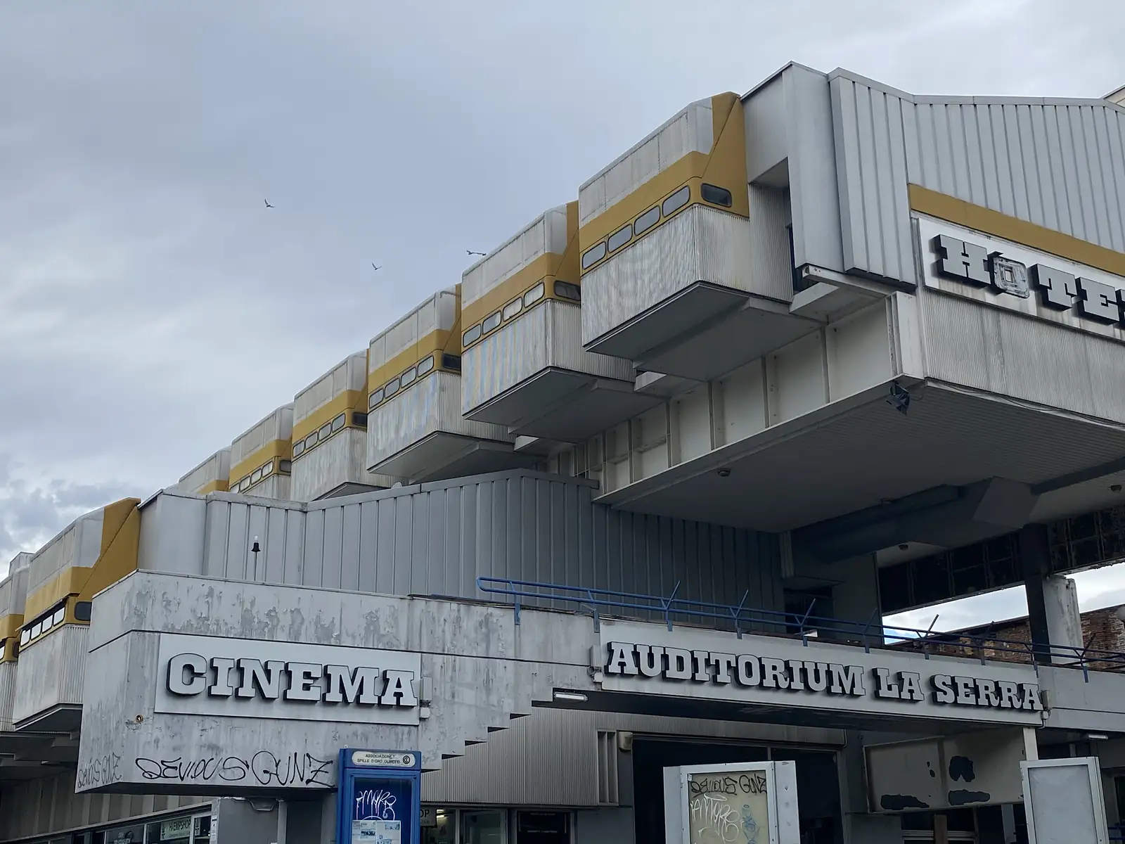 A row of grimy, yellow-striped guest-room pods projecting from the abandoned Hotel La Serra building, seen from below against a cloudy sky.