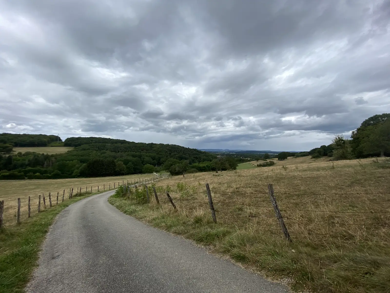 A narrow lane winds between barbed-wire pasture fences across rolling farmland under a heavy, storm-streaked sky.
