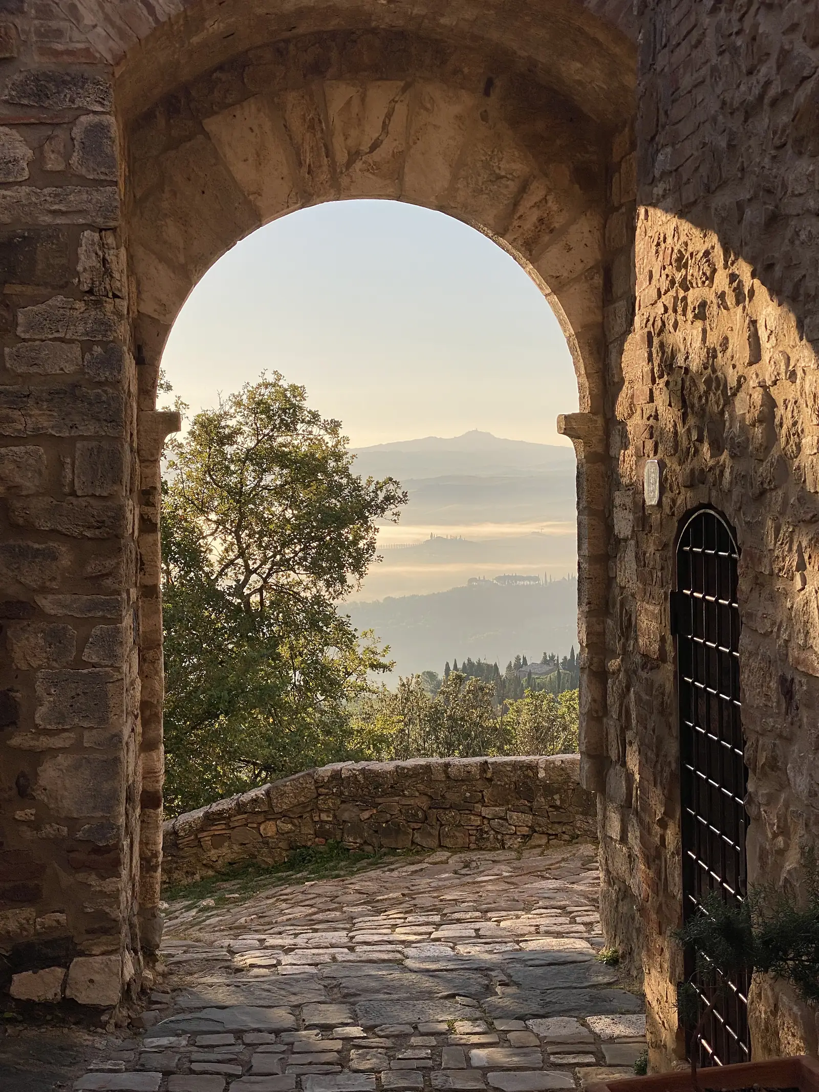 An arched stone gateway framing a hazy sunrise view over wooded hills, with an iron gate visible above the opening.