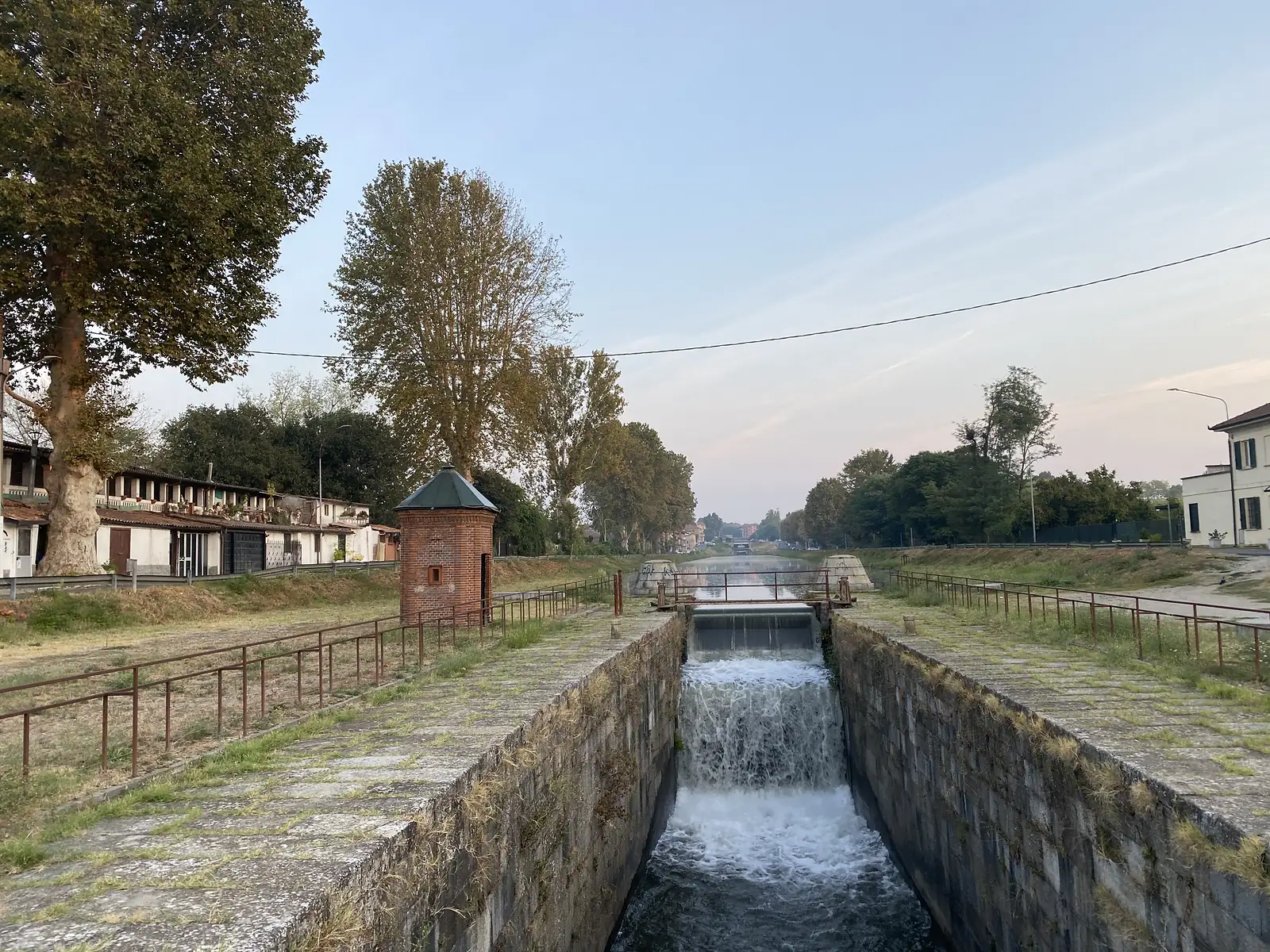 Water cascades down a stepped stone canal lock past a round brick sentry box, with houses and trees lining the towpath beyond.