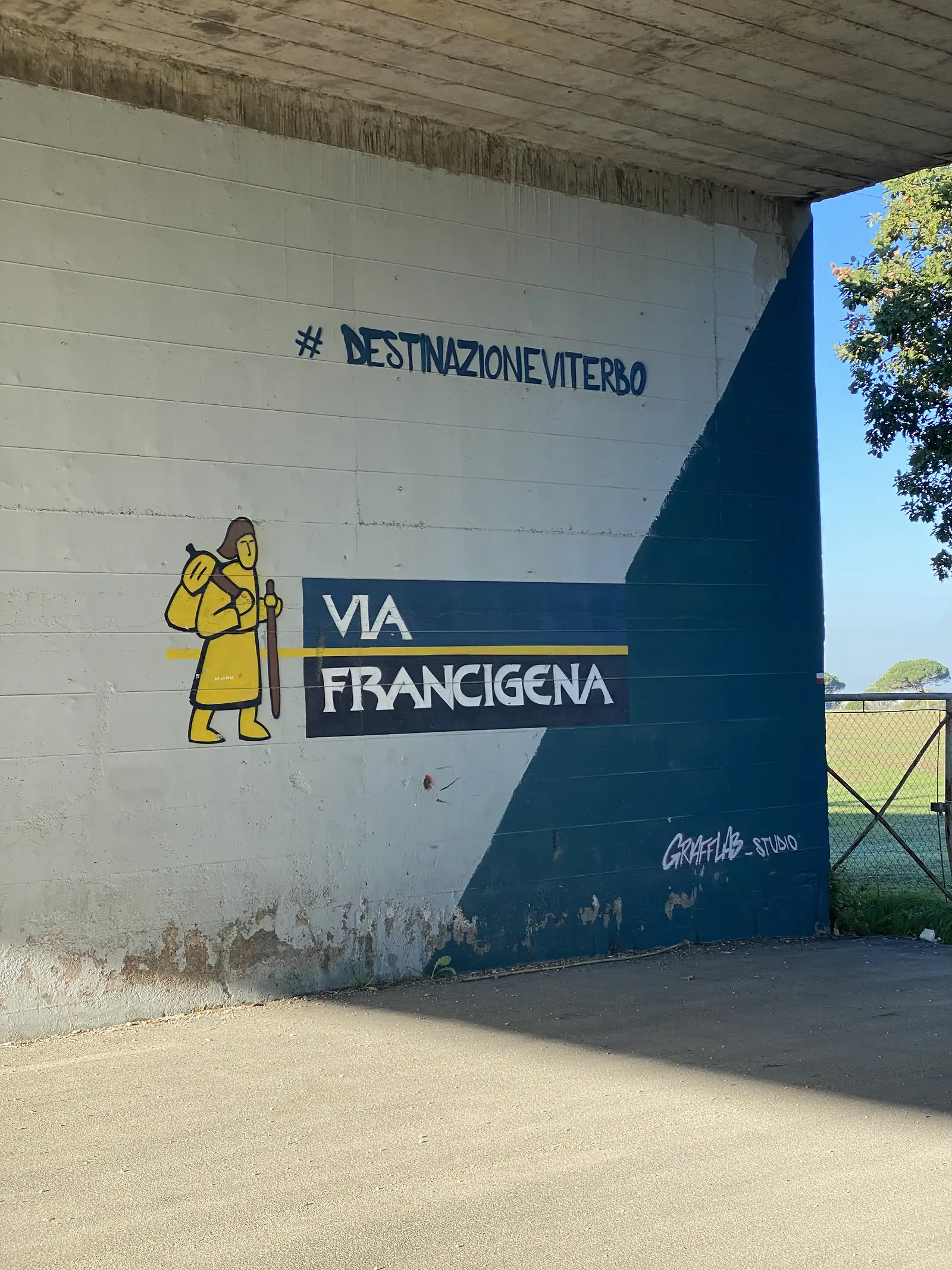 A mural on an underpass wall shows a cartoon pilgrim carrying a stick and bundle beside the words "VIA FRANCIGENA" and the hashtag "#DESTINAZIONEVITERBO".