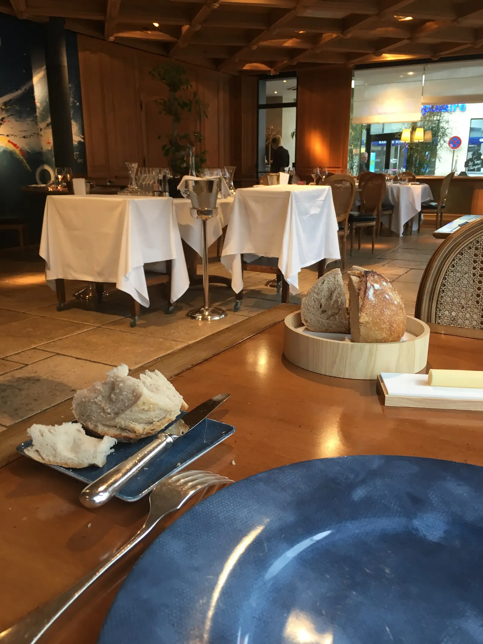 A crusty round loaf of bread in a wooden basket beside a place setting with a torn piece of bread on a blue plate, in a wood-panelled restaurant dining room.