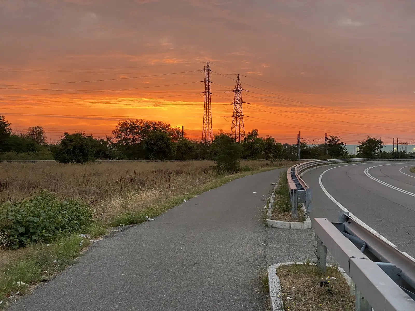 A tarmac cycle path curving beside a guardrail-lined road as pylons stand silhouetted against a fiery orange sunset.