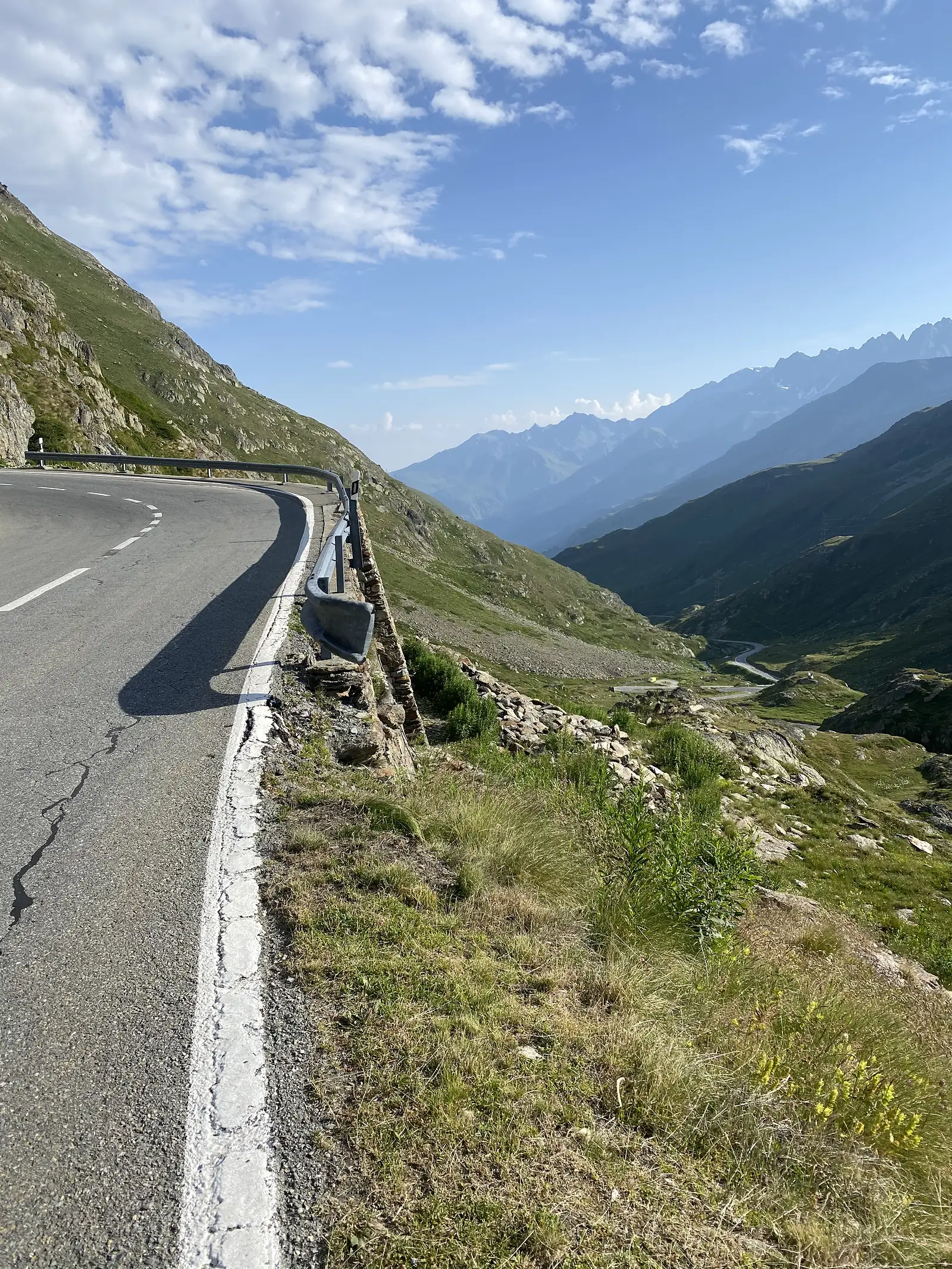 A mountain road follows a guardrail along a rocky ledge, overlooking a deep green valley and distant peaks.