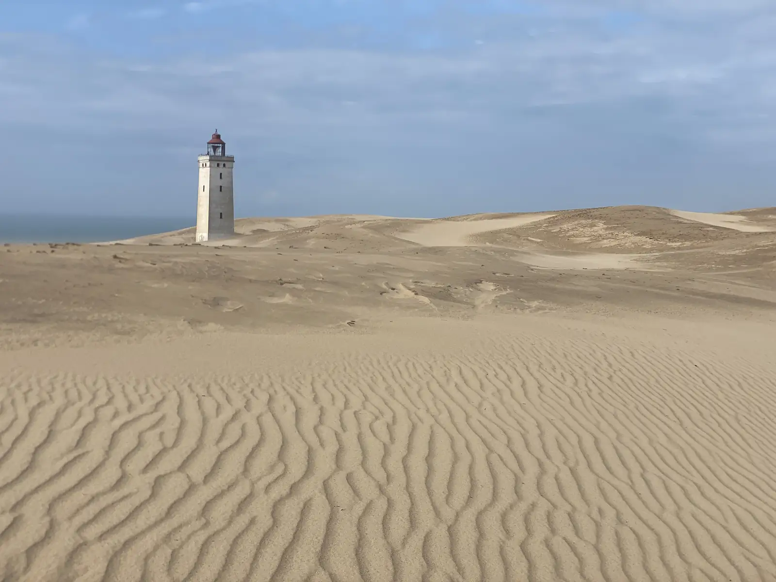 A white lighthouse with a red lantern standing above a vast rippled sand dune, with the sea beyond.