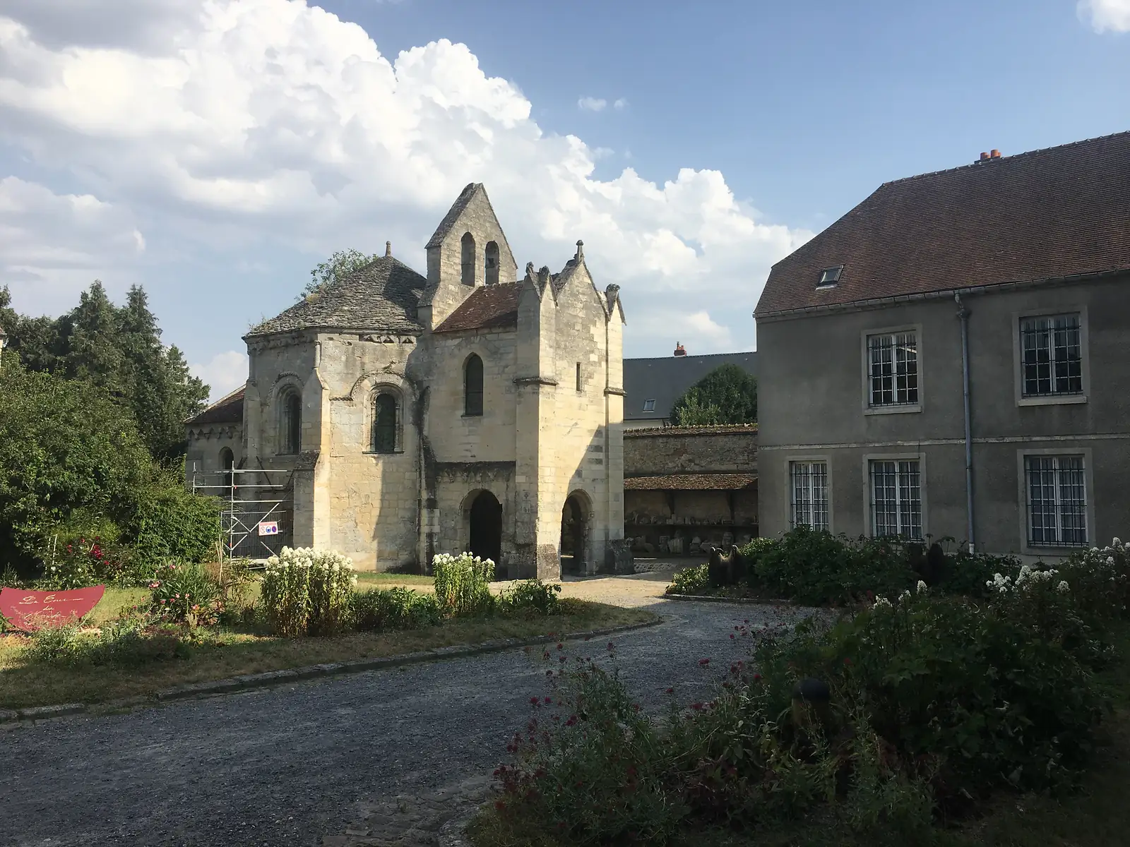 A small Romanesque chapel with an octagonal apse and a pyramidal stone roof stands in a garden beside a larger grey-roofed building.
