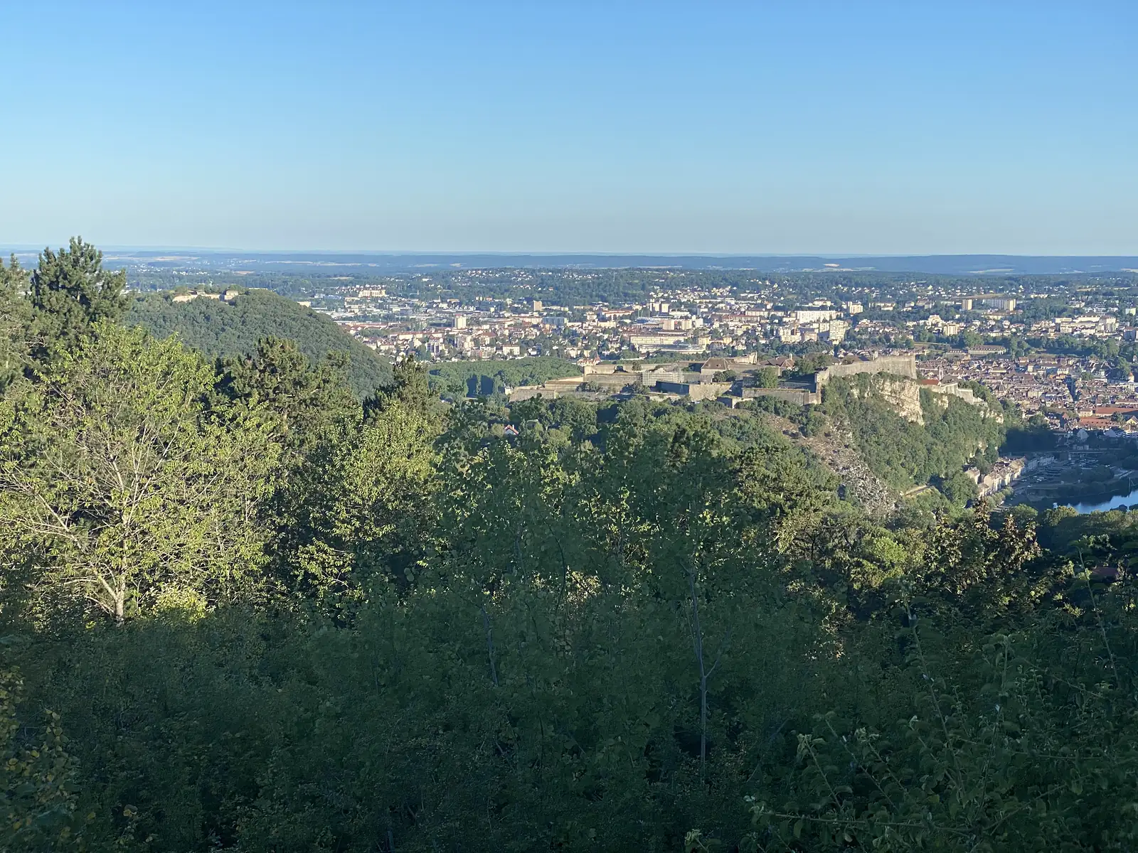 A hilltop citadel with star-shaped ramparts overlooks a river town spread across the valley floor, seen through gaps in the trees on a clear morning.