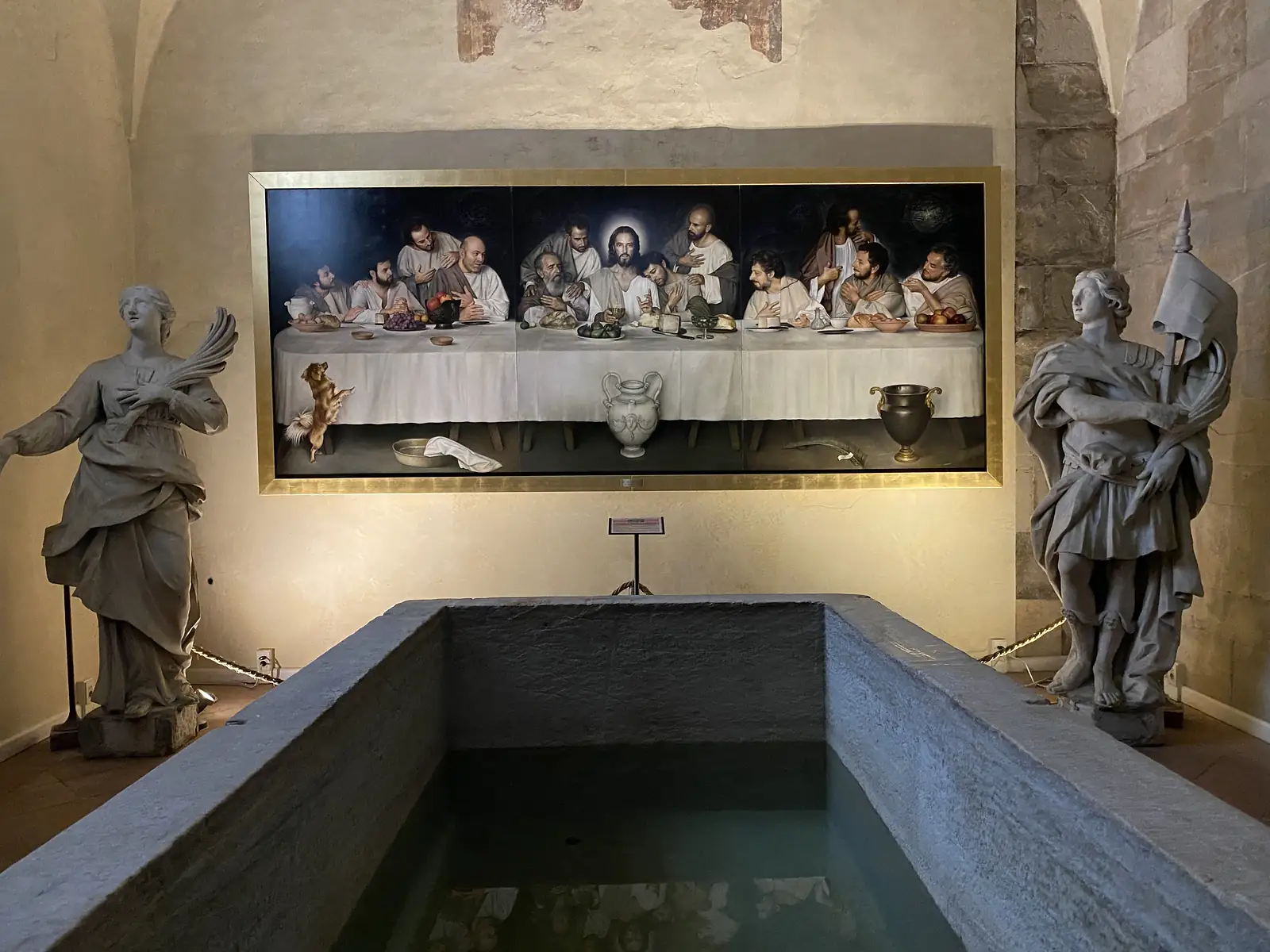 A stone baptismal pool set into a church floor, flanked by statues of saints, beneath a large painting of the Last Supper on the wall behind.