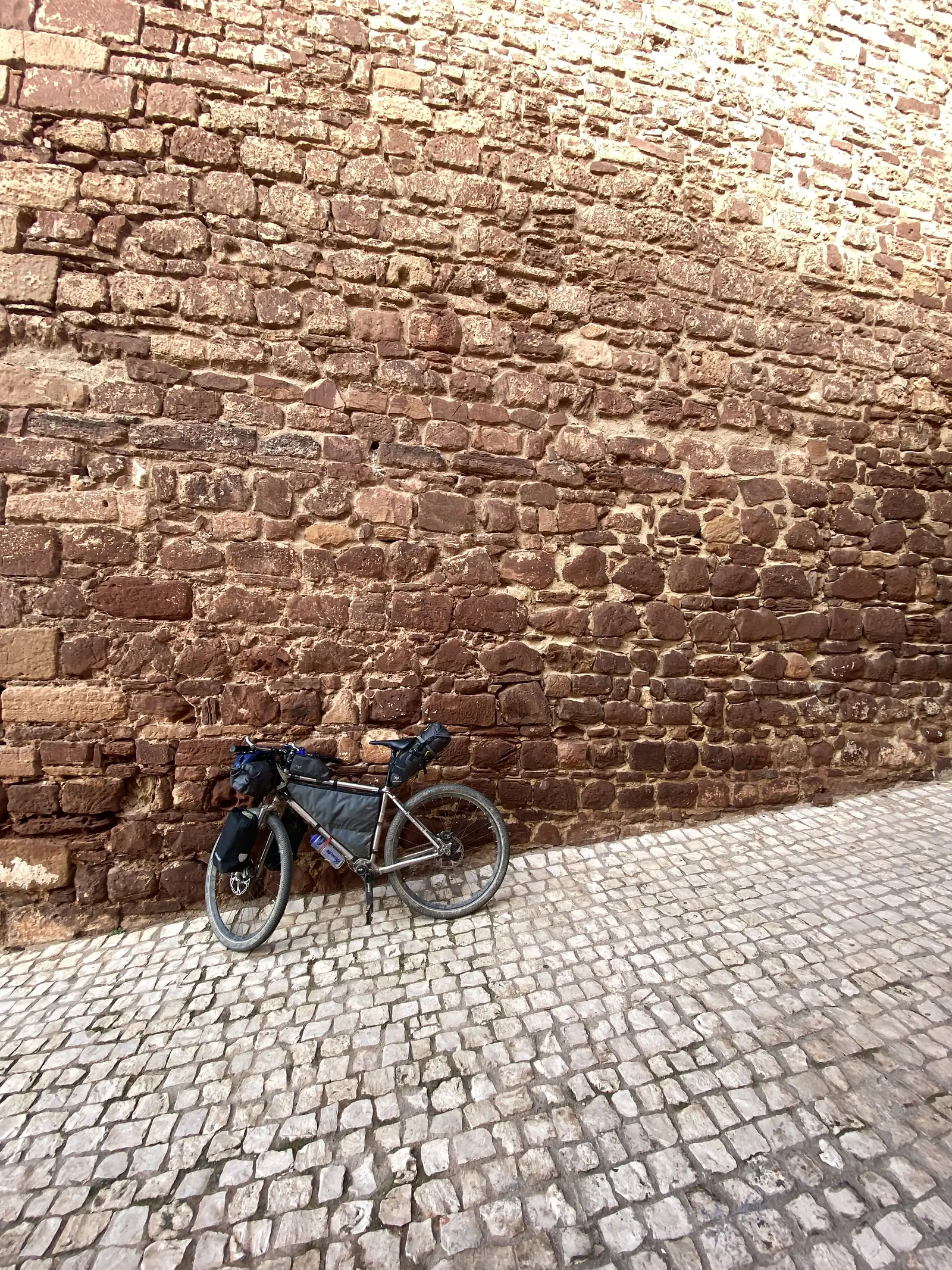 Loaded touring bike propped against the foot of a massive red sandstone castle wall, dwarfed by the stonework, on a pale cobbled apron.