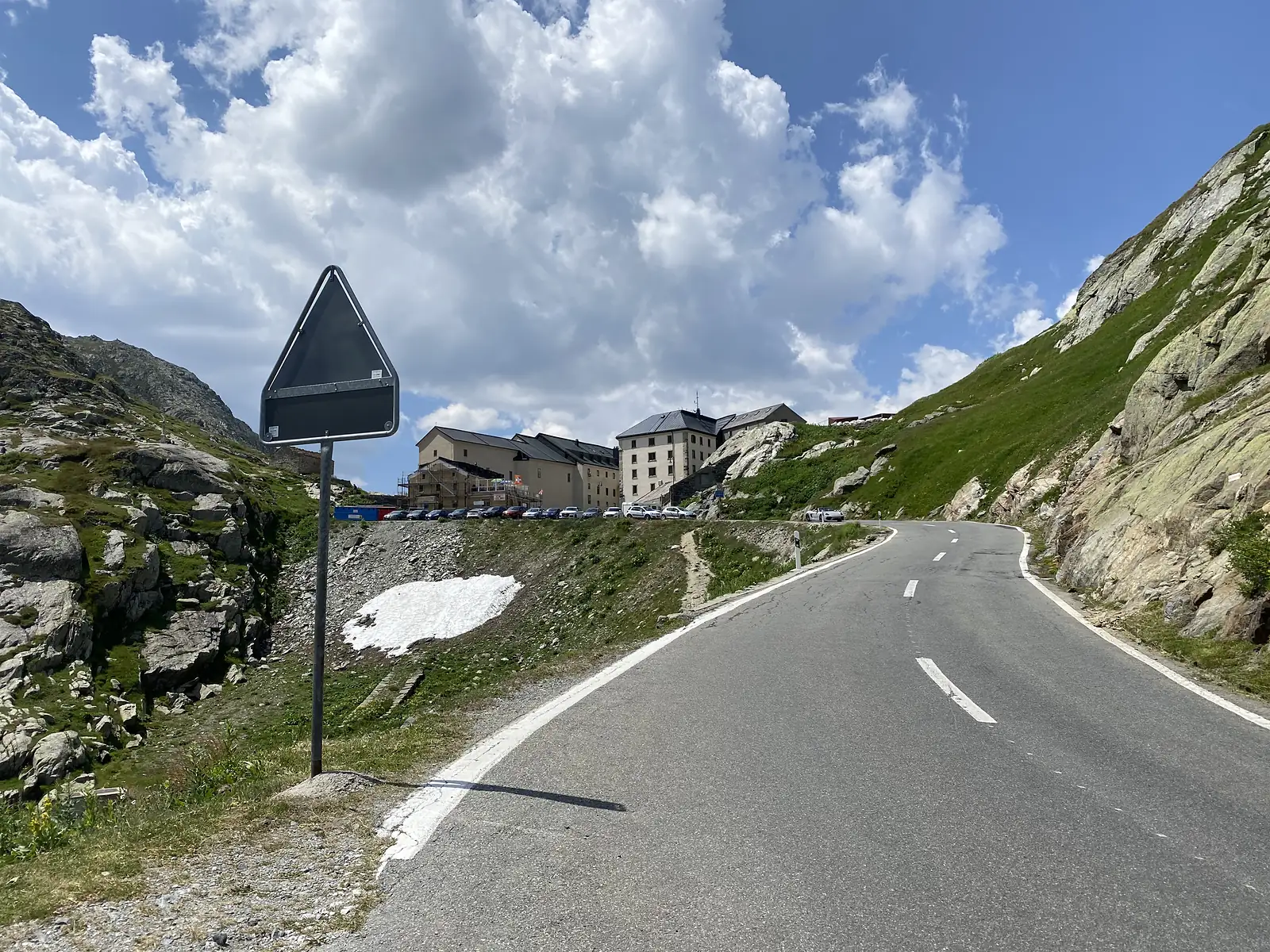 
The back of a blank triangular road sign beside the pass road, with the hospice buildings and a patch of old snow on the slope behind.
