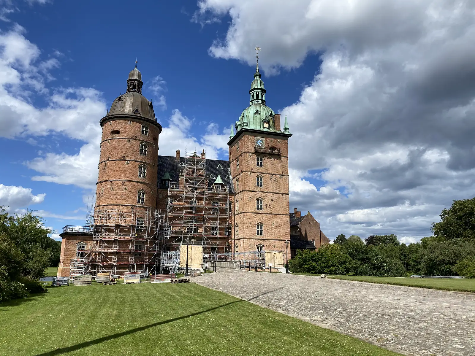 A red-brick Renaissance castle with a copper-domed round tower and a green spired clock tower, half wrapped in scaffolding above a cobbled drive.