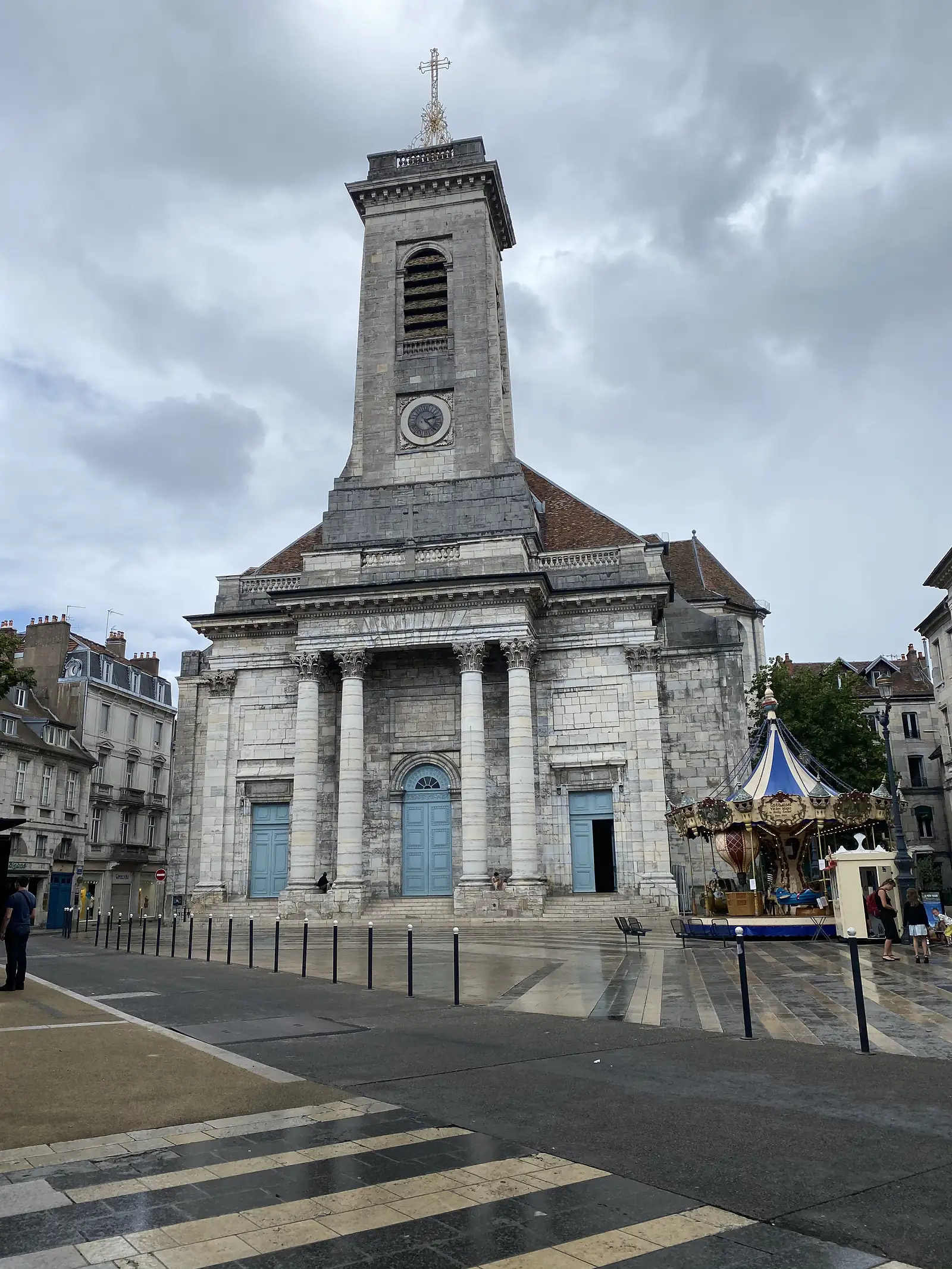 The classical stone facade and tall clock tower of a Besançon church rise above a paved square where a fairground carousel stands to one side.