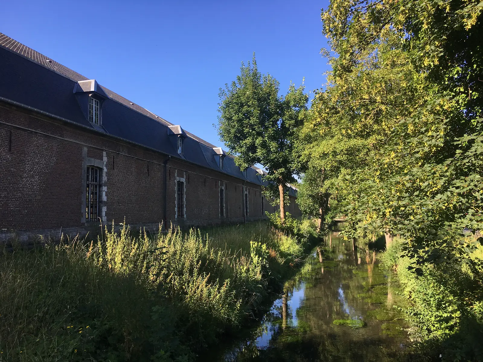 A long brick citadel building beside a tree-lined moat reflecting the sky, part of Arras's star-shaped fortifications.