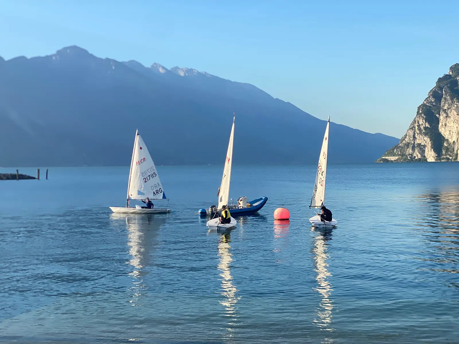 Three single-handed dinghies with white sails drift on flat water beside an orange marker buoy and a coach's inflatable, with a long mountain ridge behind.