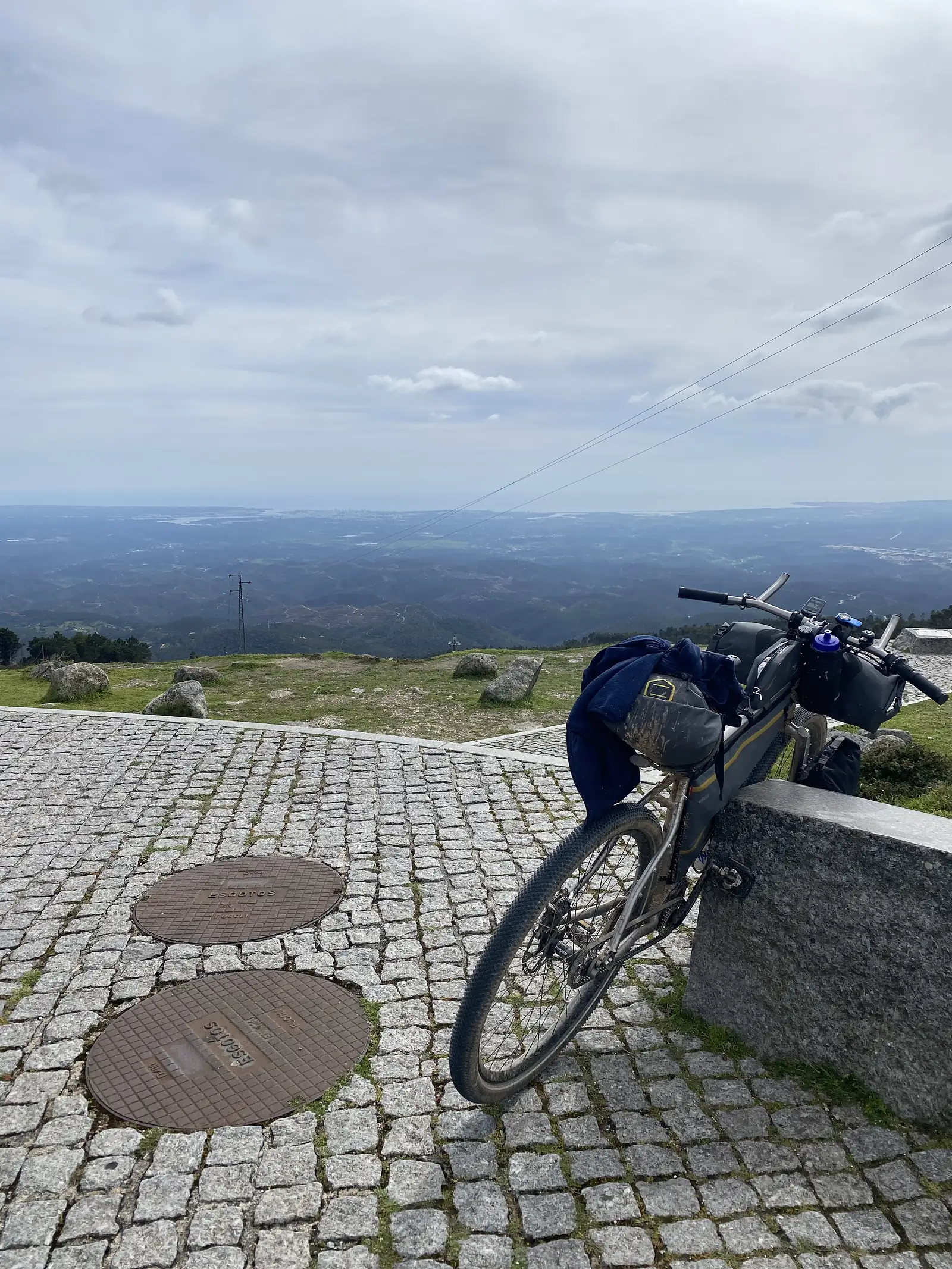 Loaded bike leaning against a granite block on a cobbled hilltop viewpoint, a jacket slung over the bars, with wooded hills falling away to a distant hazy coastline.