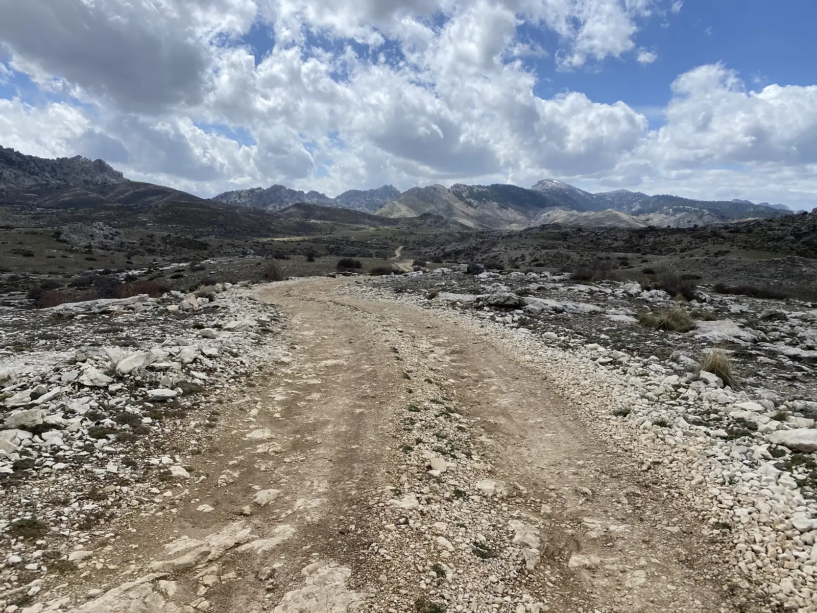 A stony track running down towards a bowl of pale limestone hills and jagged peaks, one streaked with snow.