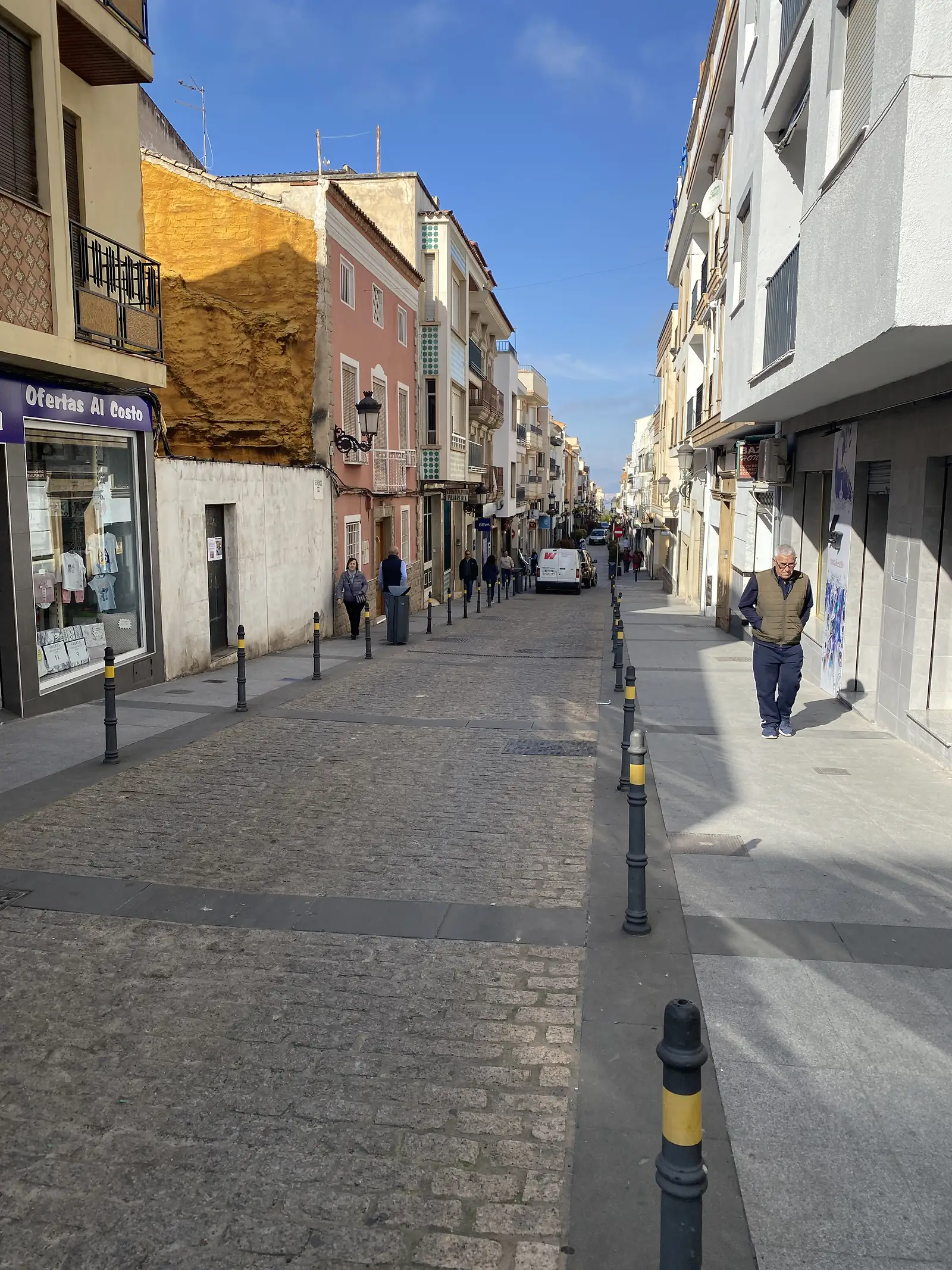 A cobbled pedestrian street lined with small shops and bollards, a man walking towards the camera in the morning sun.