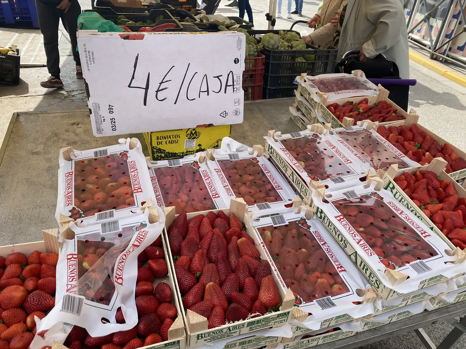 A market stall of strawberries in wooden crates behind a hand-written cardboard sign reading "4 euros a box".