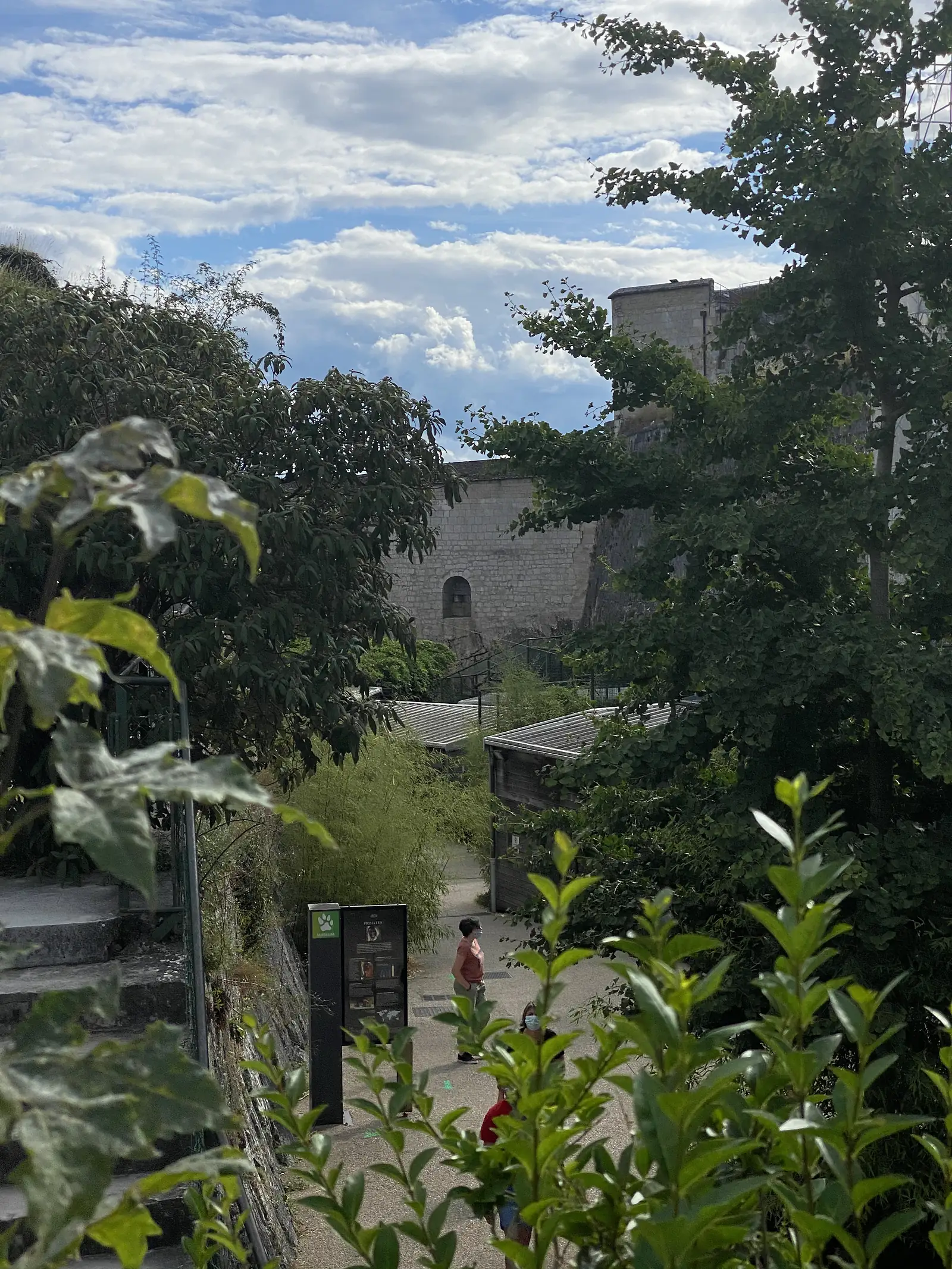 A gravel path lined with trees and shrubs leads down past a signpost towards enclosures in the citadel's zoo grounds, with visitors walking ahead.