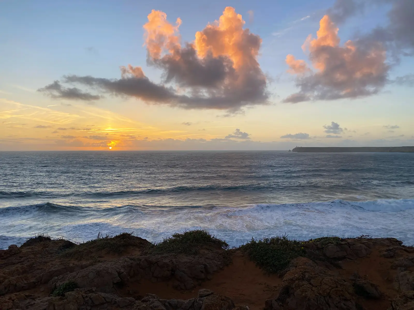 Sun dropping to the sea horizon beneath pink-lit cumulus, waves breaking in ranks onto a rocky red foreshore with a long headland silhouetted to the right.