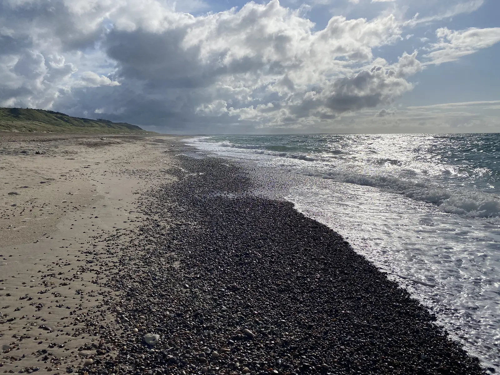 A long strand where a broad band of dark pebbles meets pale sand and breaking waves, with dune cliffs receding into the distance.
