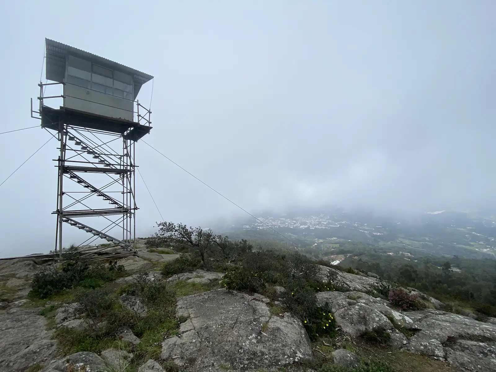 Glass-sided fire lookout tower on steel scaffolding legs, guyed with wires, standing on a rocky summit above a valley town that shows through a break in the mist.
