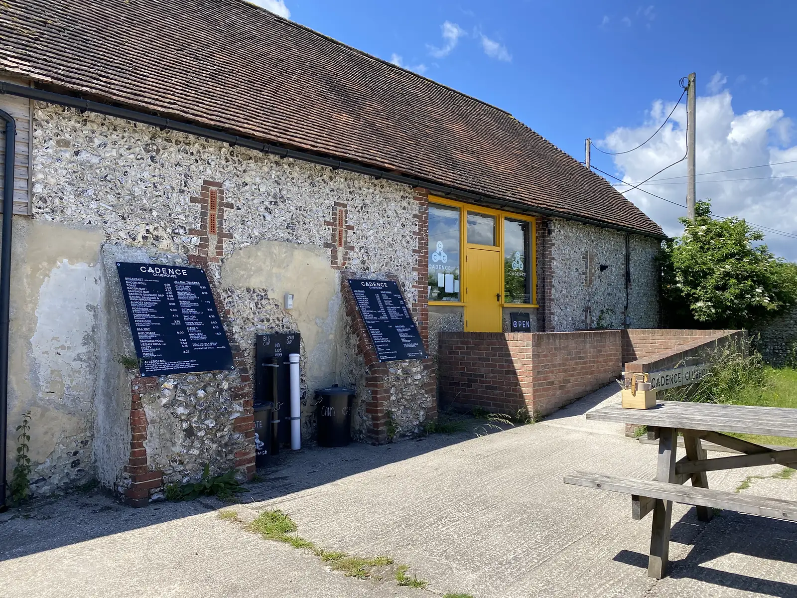 A flint and brick barn converted to a cyclists' cafe, lettered "CADENCE CLUBHOUSE" with a yellow door and menu boards outside.