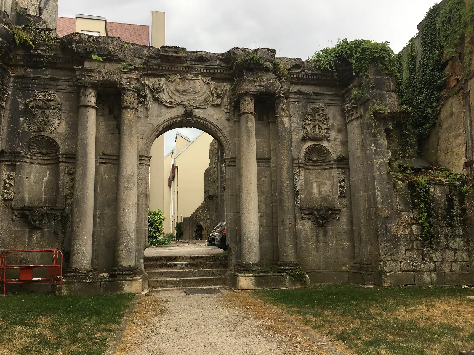 The ruined stone facade of a former church in Langres, its columned Baroque archway open to the sky with ivy climbing the surviving wall and parked cars visible through the arch.